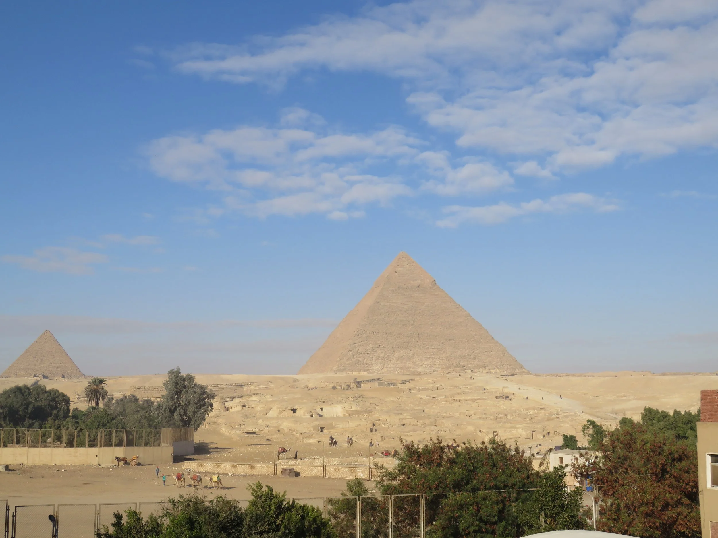 View of the Great Pyramid of Giza under a partly cloudy sky, with some greenery and small buildings in the foreground.