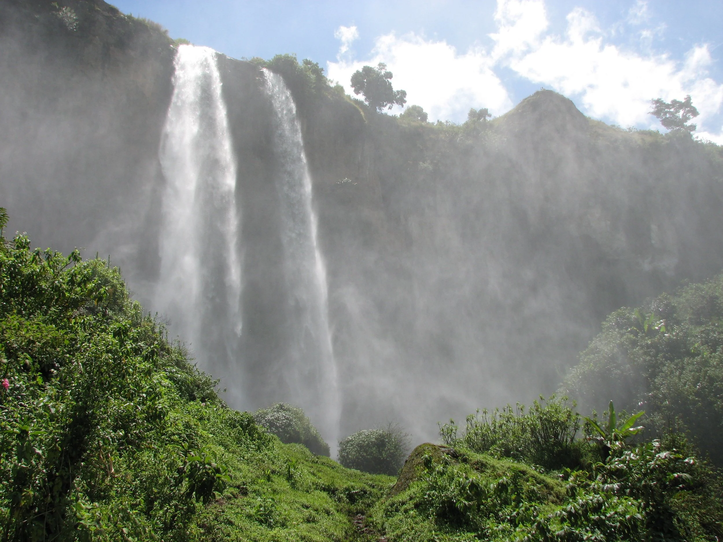 Large waterfall cascading down a mountain surrounded by lush green vegetation under a blue sky with some clouds.