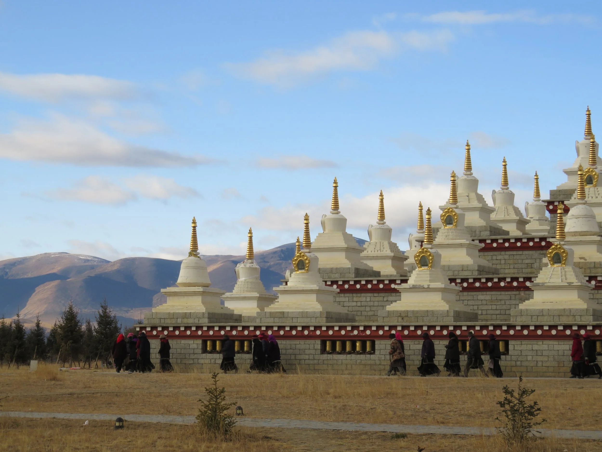 A group of people walking near white and gold stupas in a field with mountains and blue sky in the background.