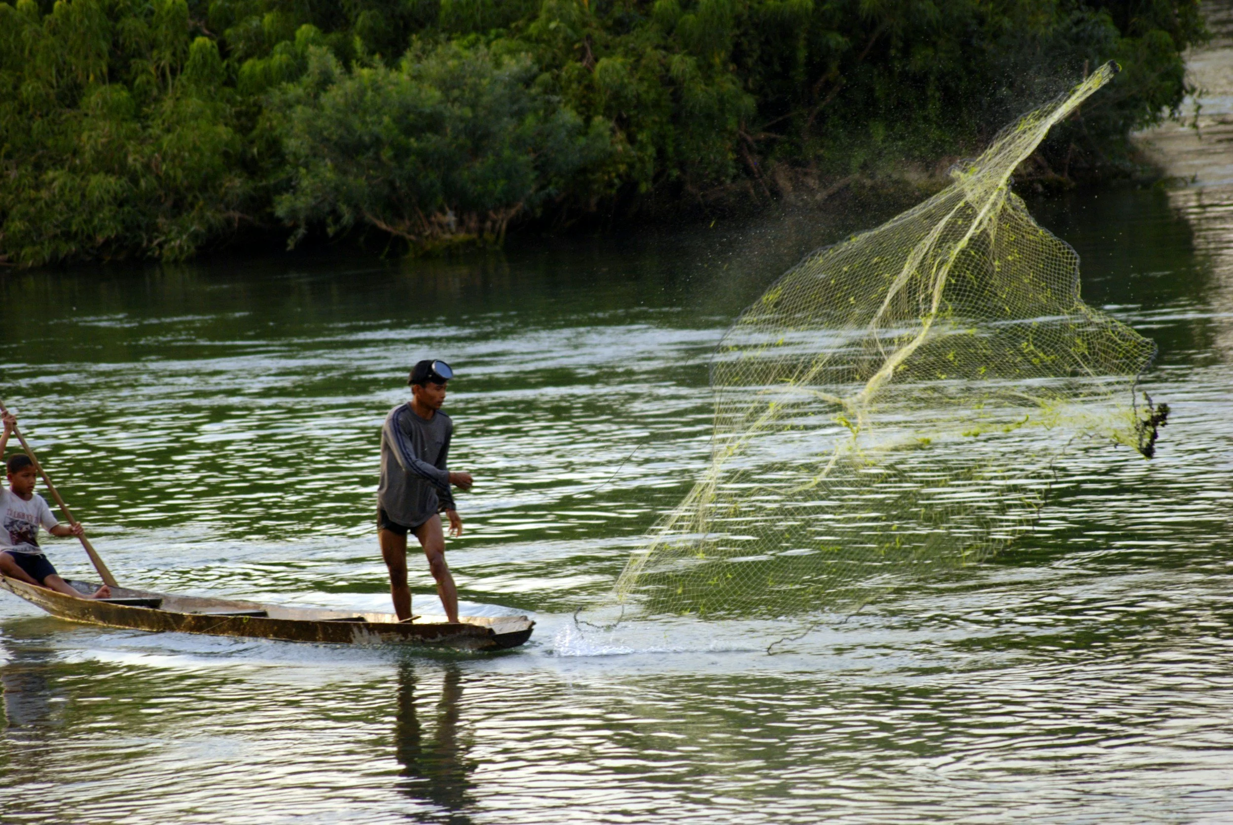 Two children on a wooden boat in a river, one standing and casting a fishing net, the other sitting and holding a pole, with green trees in the background.