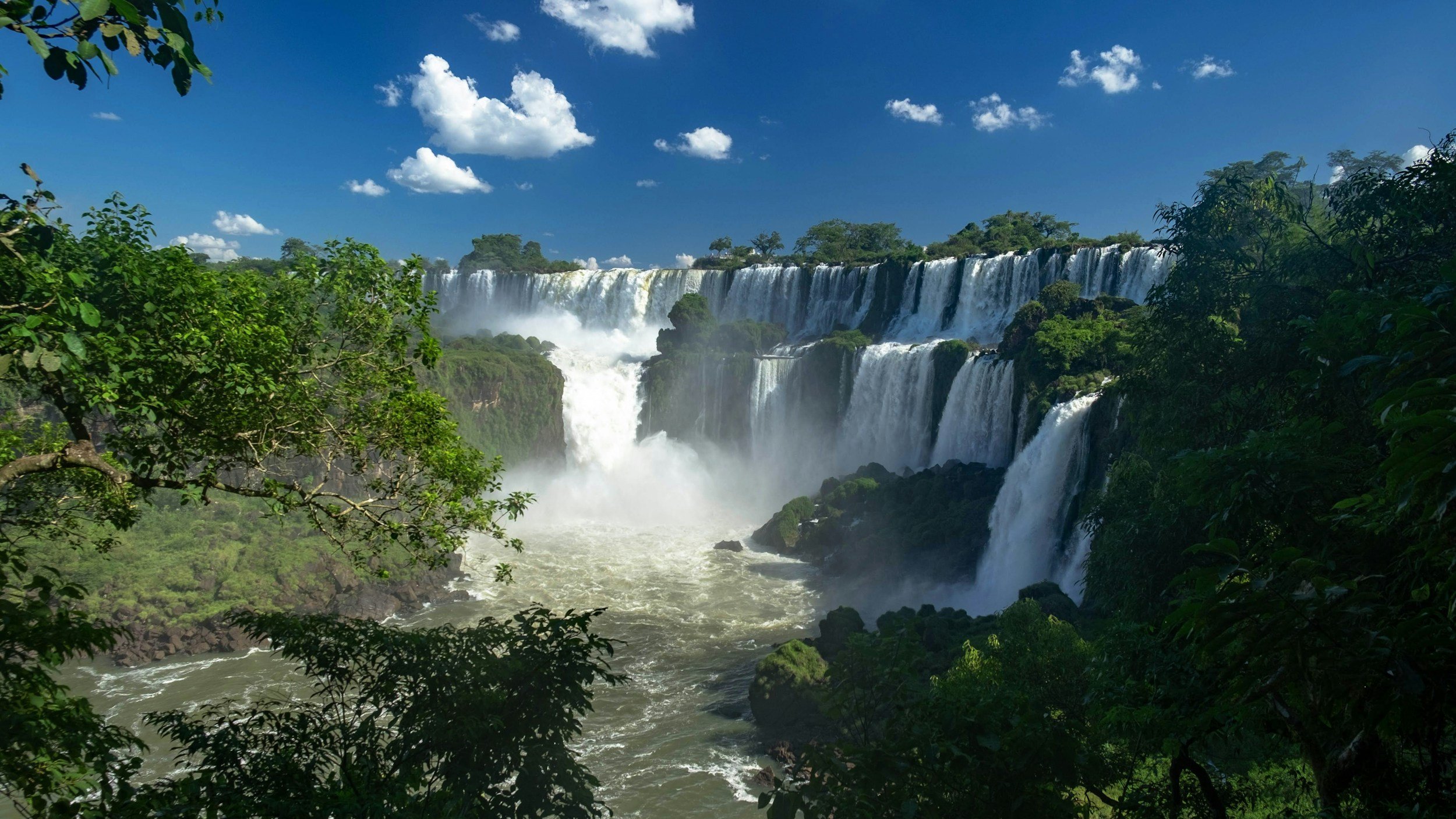 A scenic view of Iguazu Falls with cascading waterfalls surrounded by lush green trees under a bright blue sky with white clouds.