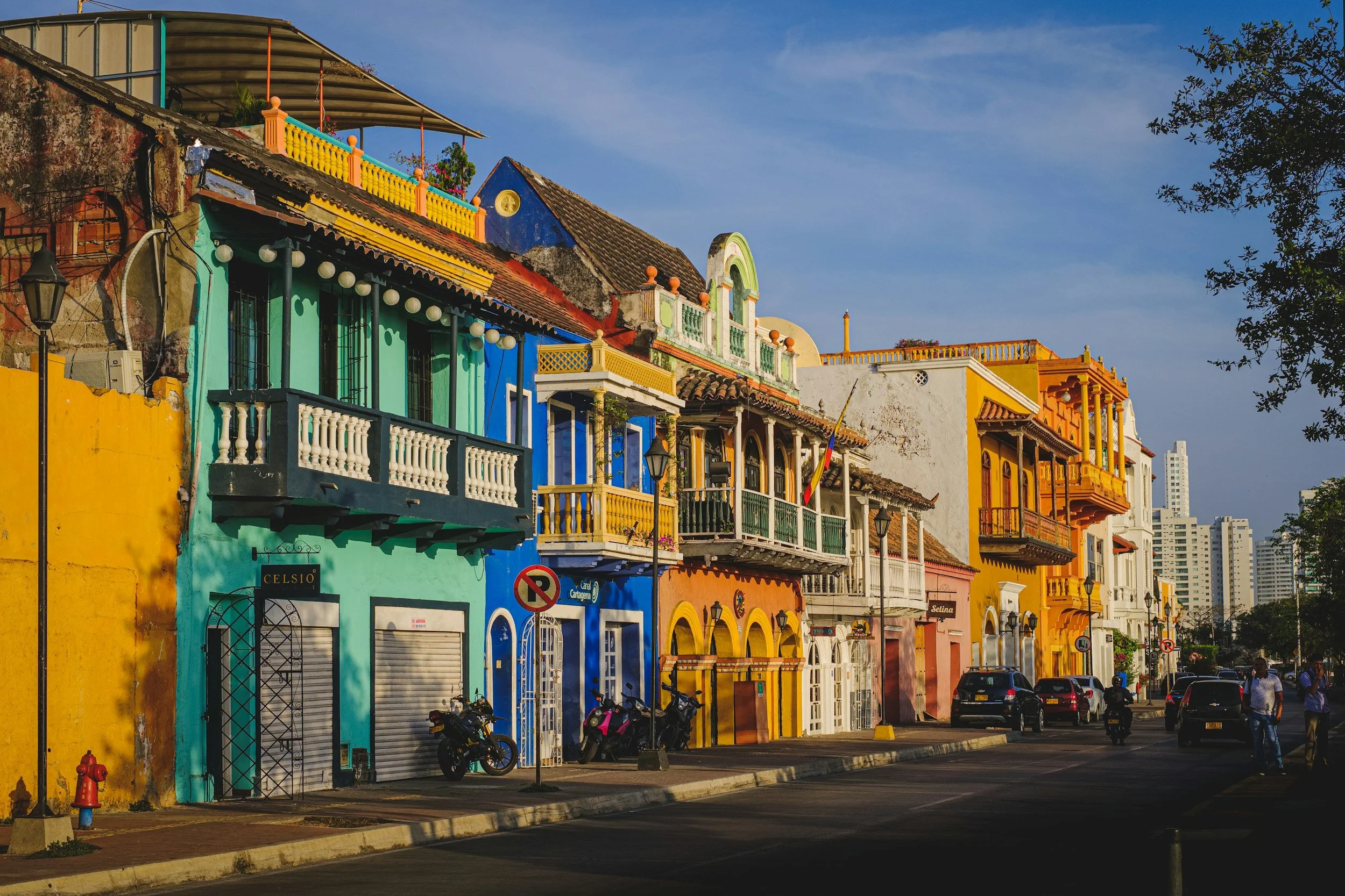 Colorful colonial-style buildings in vibrant yellow, blue, orange, and white along a city street with cars, motorcycles, and people walking, under a clear blue sky.