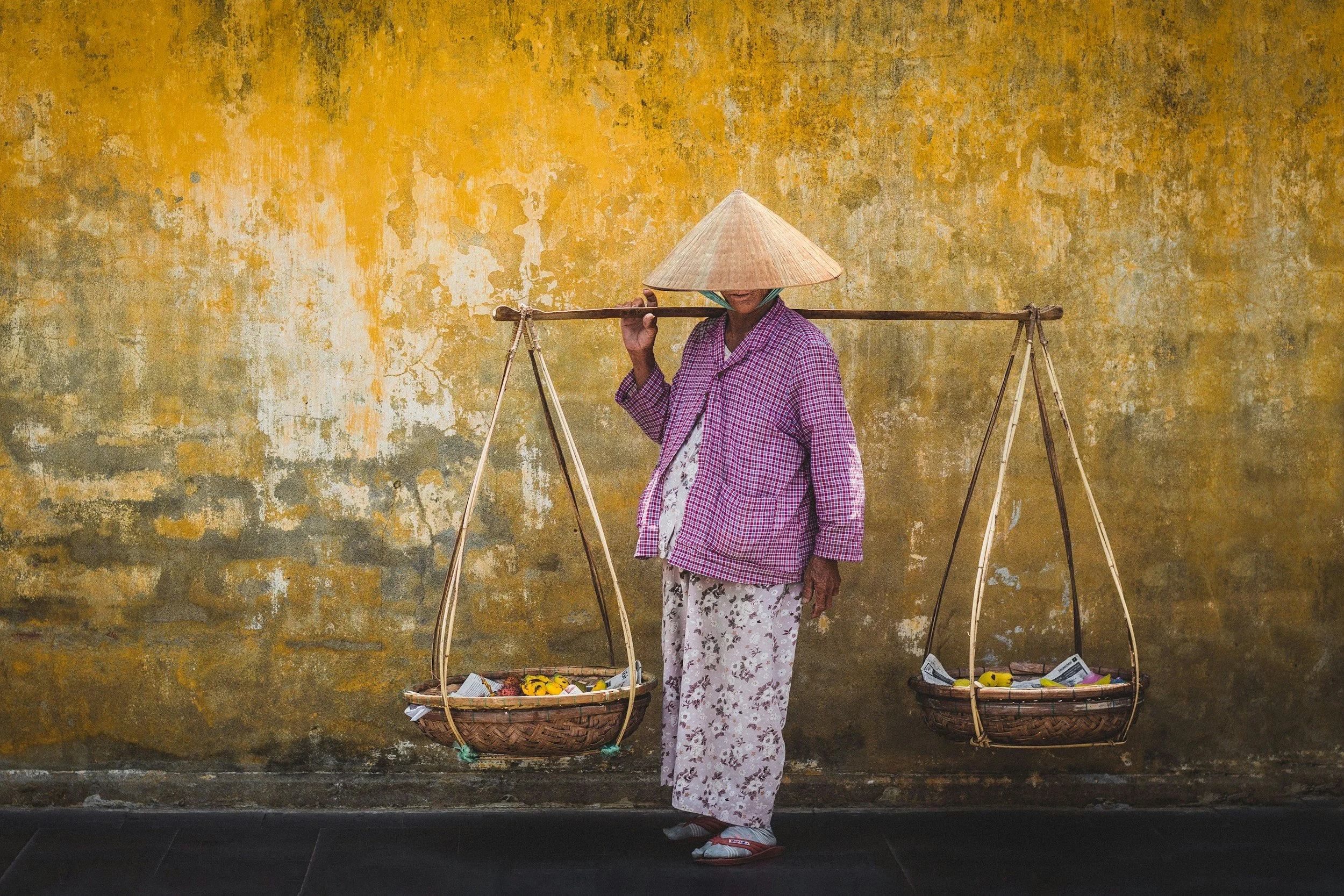 A person wearing a conical hat and patterned clothing carries a traditional bamboo pole with baskets hanging on each side, filled with flowers and paperwork, walking in front of a textured yellow wall.