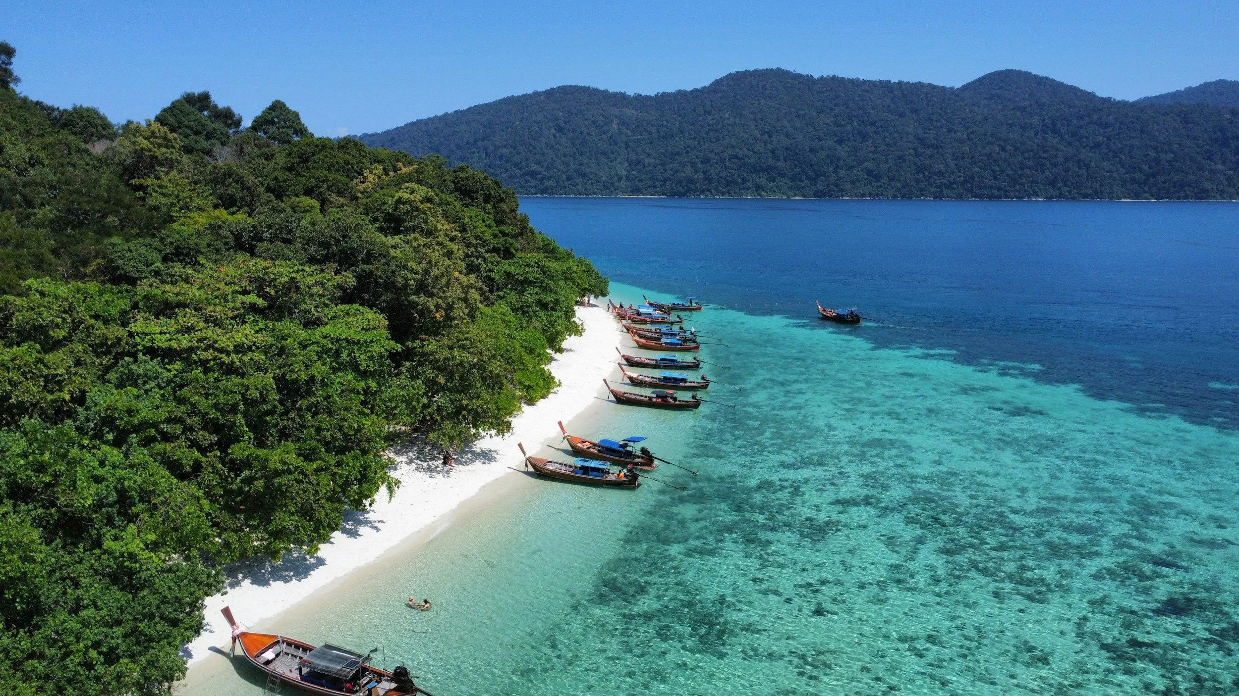 A tropical beach with white sand, several boats anchored near the shore, lush green trees on the land, and clear turquoise water, with hills and mountains in the background.