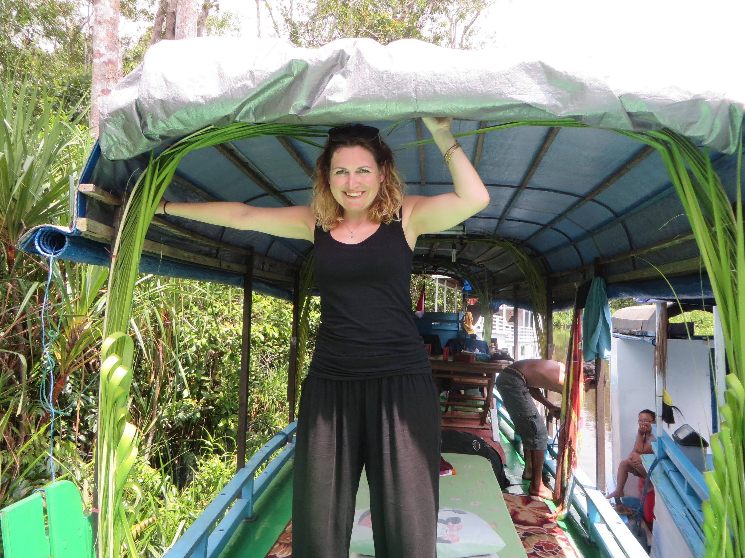 A woman in a black sleeveless top and black pants smiling while standing inside a boat with a blue and green canopy, surrounded by greenery and other people working in the background.