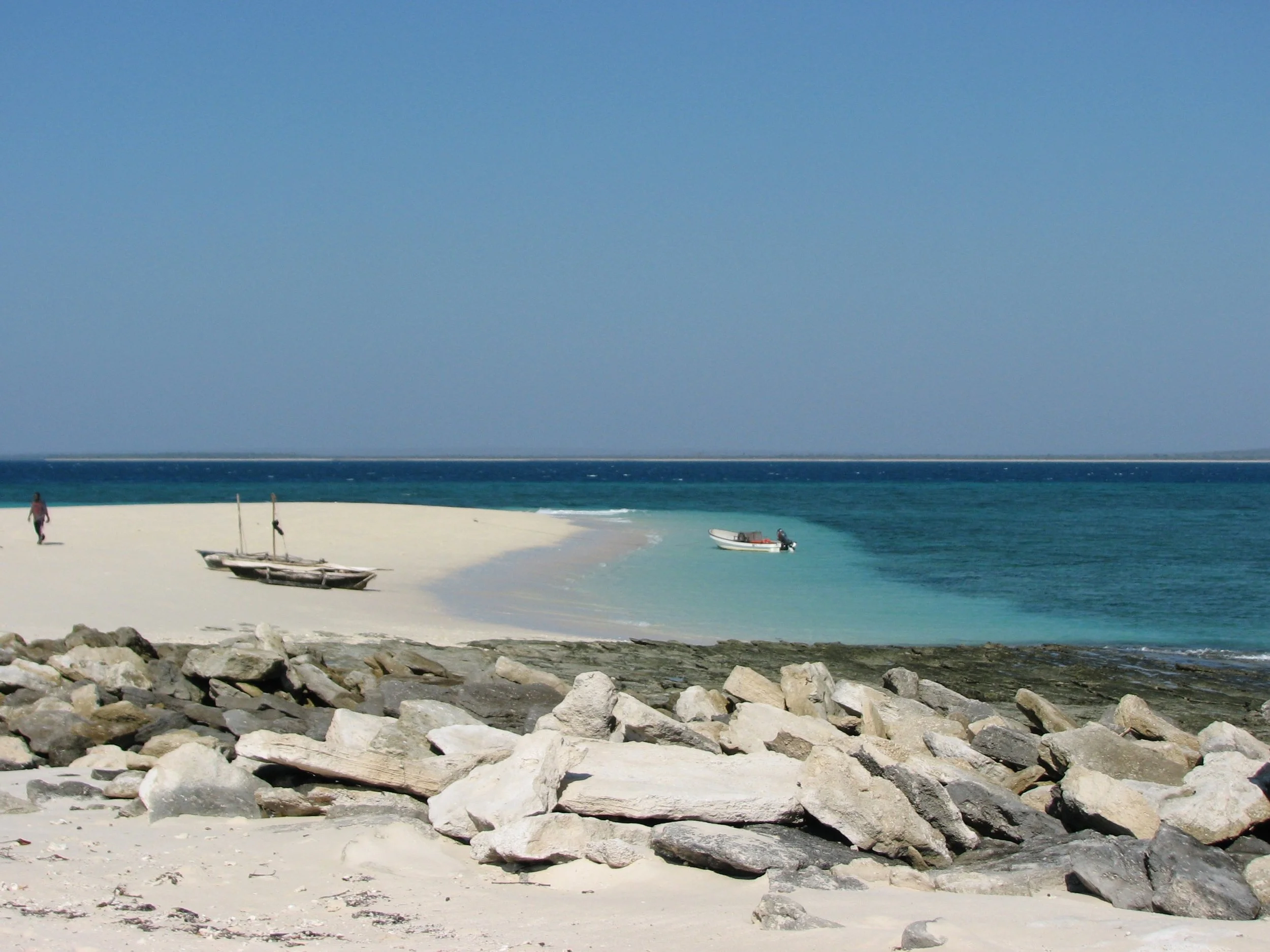 A peaceful beach scene with rocks in the foreground, white sand, two boats in the water, and a person walking along the beach under a clear blue sky.