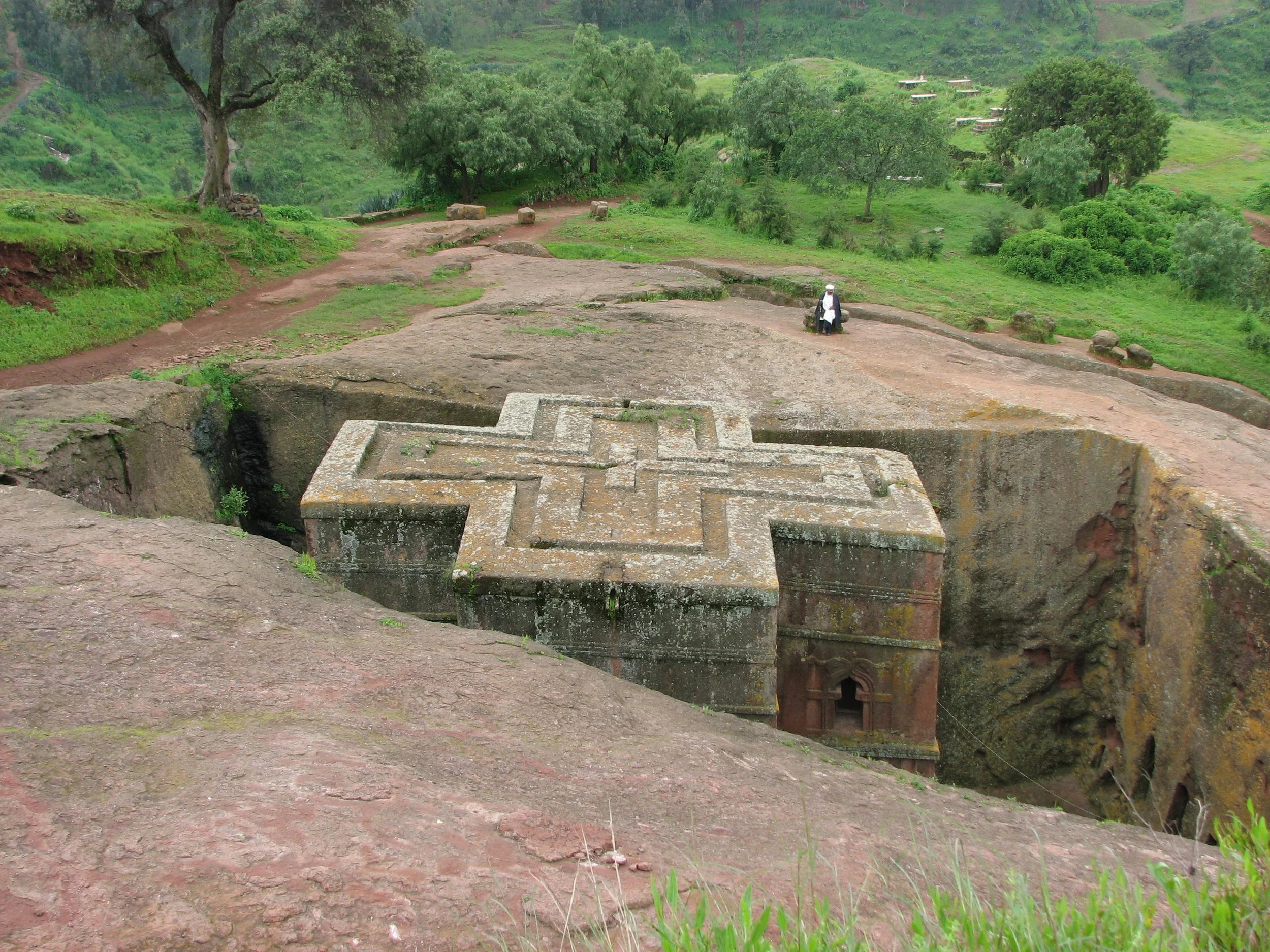 Ancient rock-hewn monastery in Tigray, Ethiopia, with large carved cross on the stone surface, surrounded by green landscape and trees.