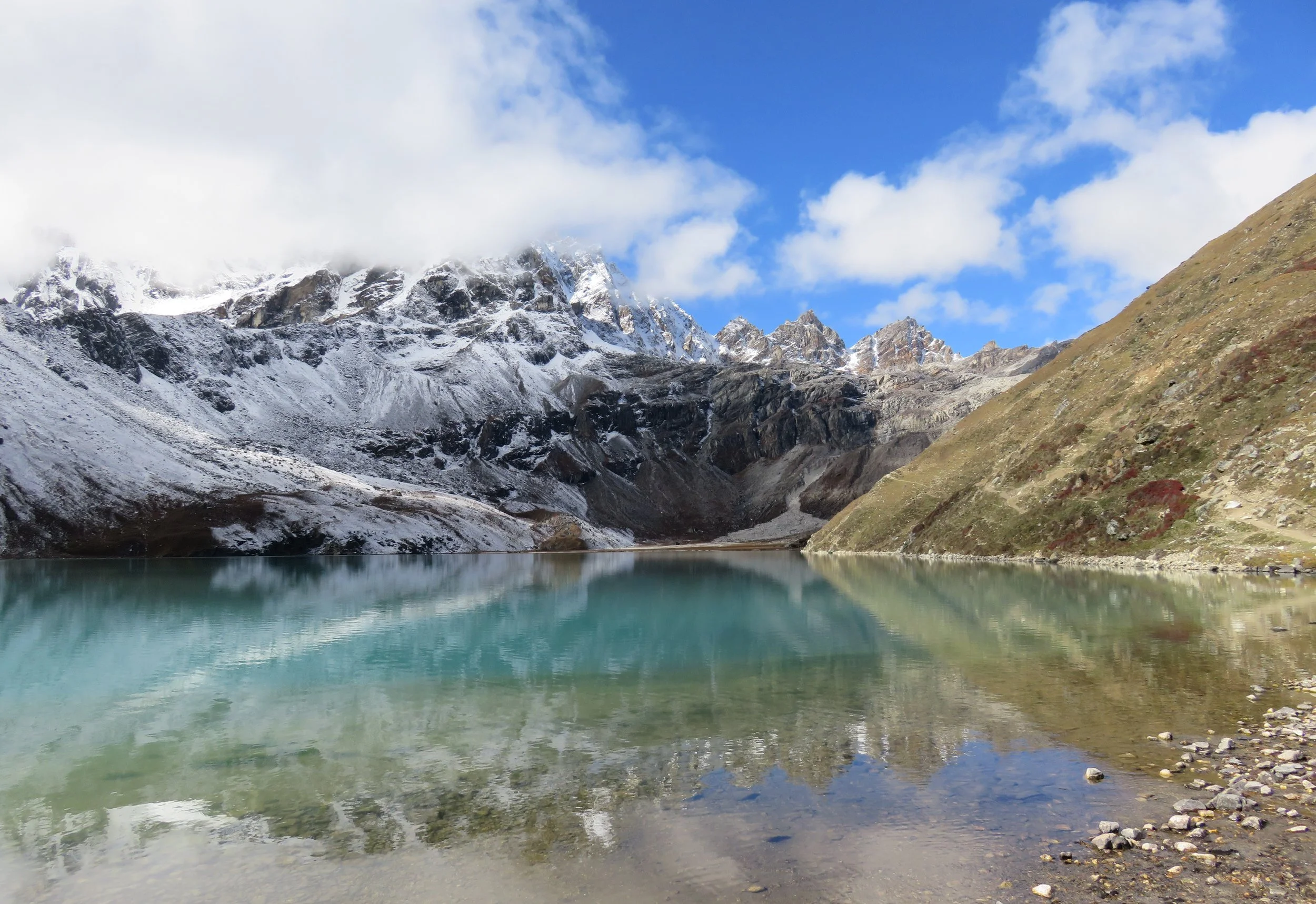 A mountain lake with clear water reflects the snow-capped mountains and blue sky with some clouds.