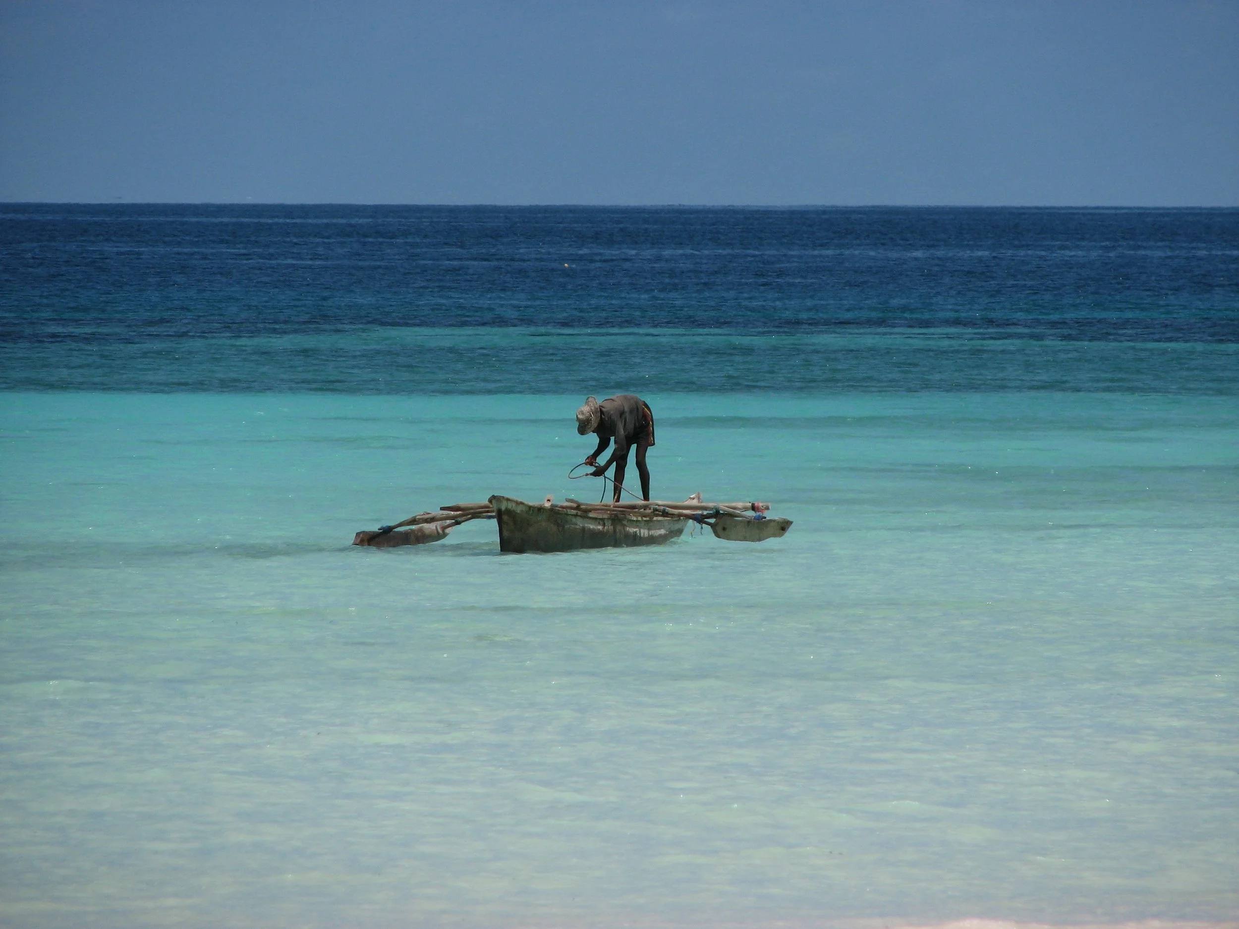 A person standing on a small boat in shallow, turquoise water near the shore, with a deep blue ocean and sky in the background.