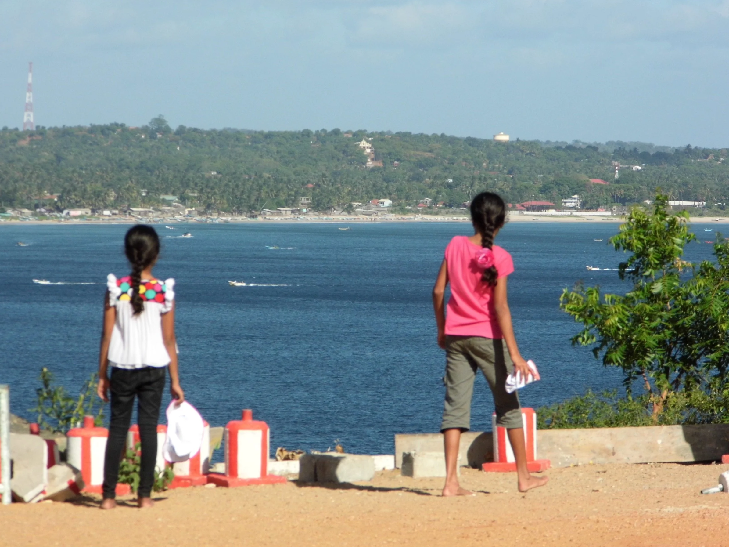 Two girls standing on a waterfront, looking at the ocean with boats passing by, and a green hillside in the background.