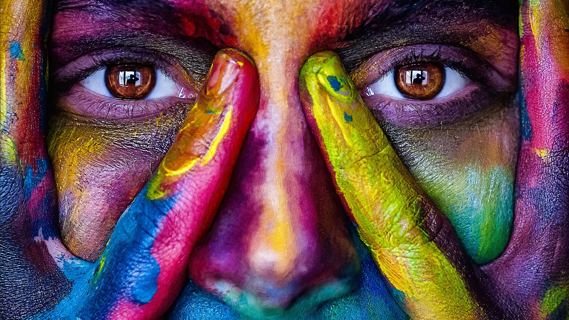Close-up of a person's face painted with vibrant, multicolored paint, with hands covering part of the face, revealing brown eyes.