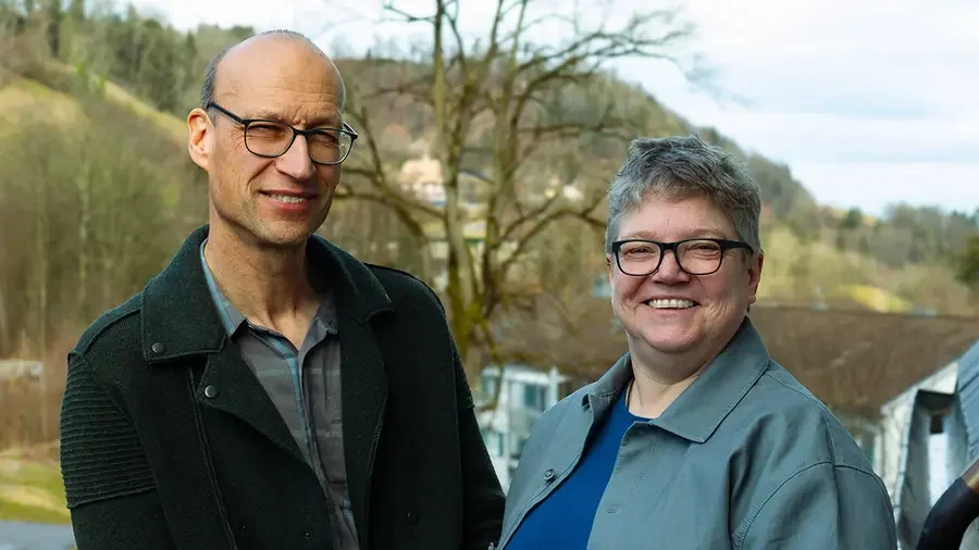 Ein Mann und eine Frau lächeln draußen vor einem ländlichen Hintergrund mit Bäumen und Häusern.