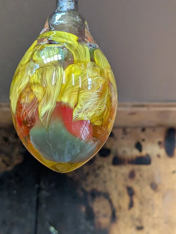Eine Glasflasche mit gelb-orangefarbenem Muster, die mit Wasser gefüllt ist, hängt an einem Faden. Im Hintergrund ein Holzblock und eine graue Wand.