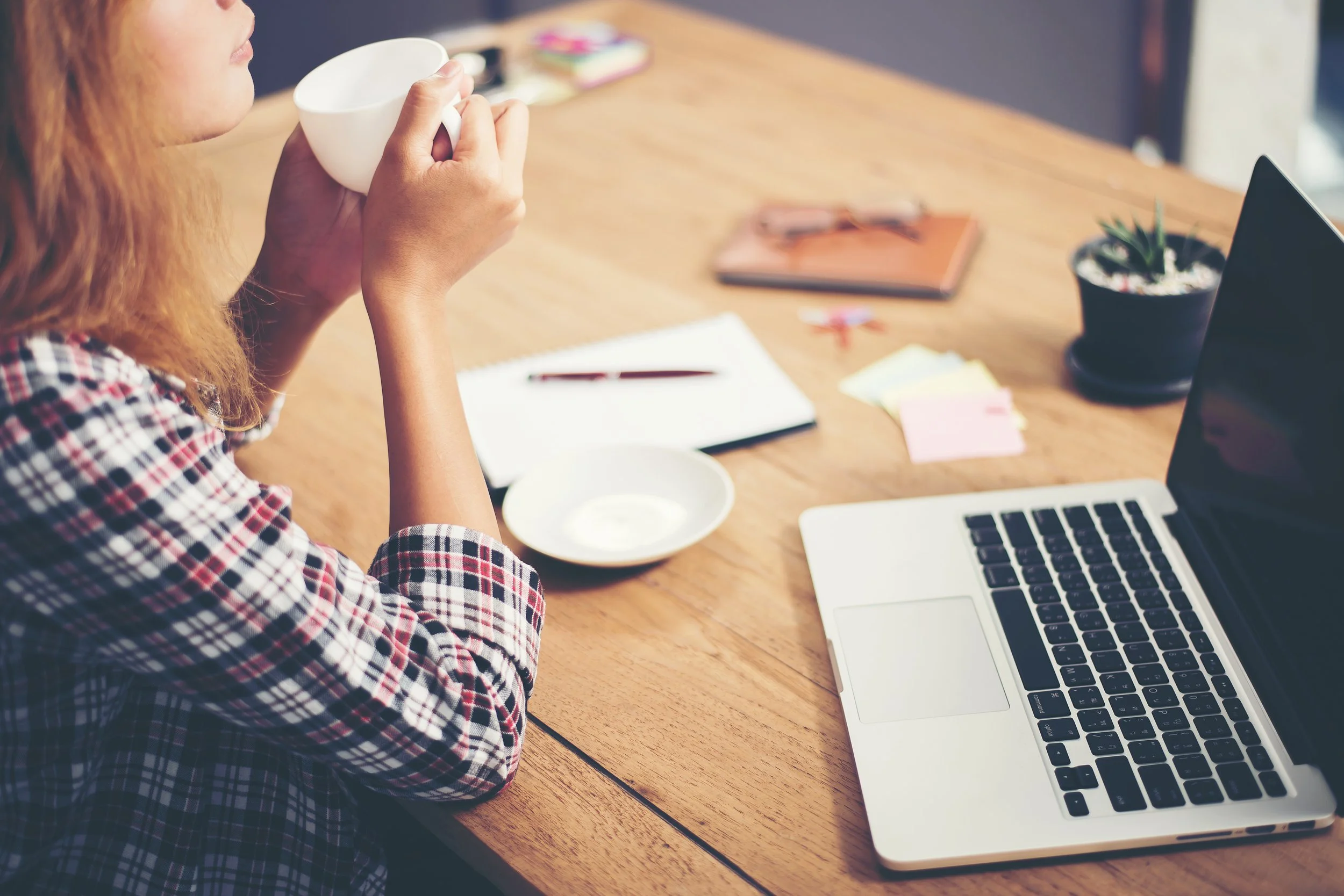 Une femme assise à un bureau en train de boire dans une tasse, avec un ordinateur portable, un carnet, des post-it, une petite plante, et des notes colorées sur la table.