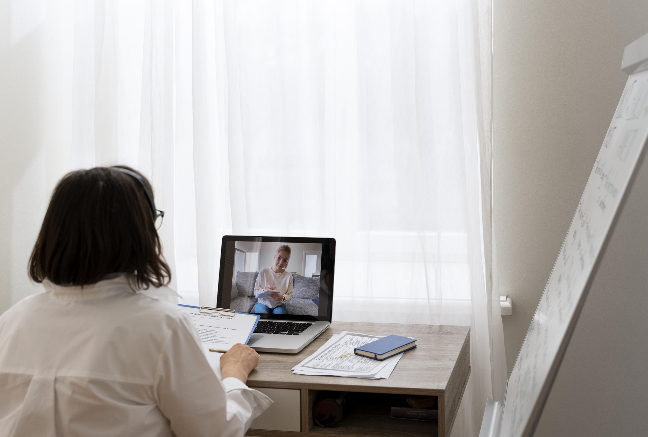 Une femme assise à son bureau lors d'une réunion virtuelle sur son ordinateur portable  avec des documents et un carnet, dans une pièce lumineuse.
