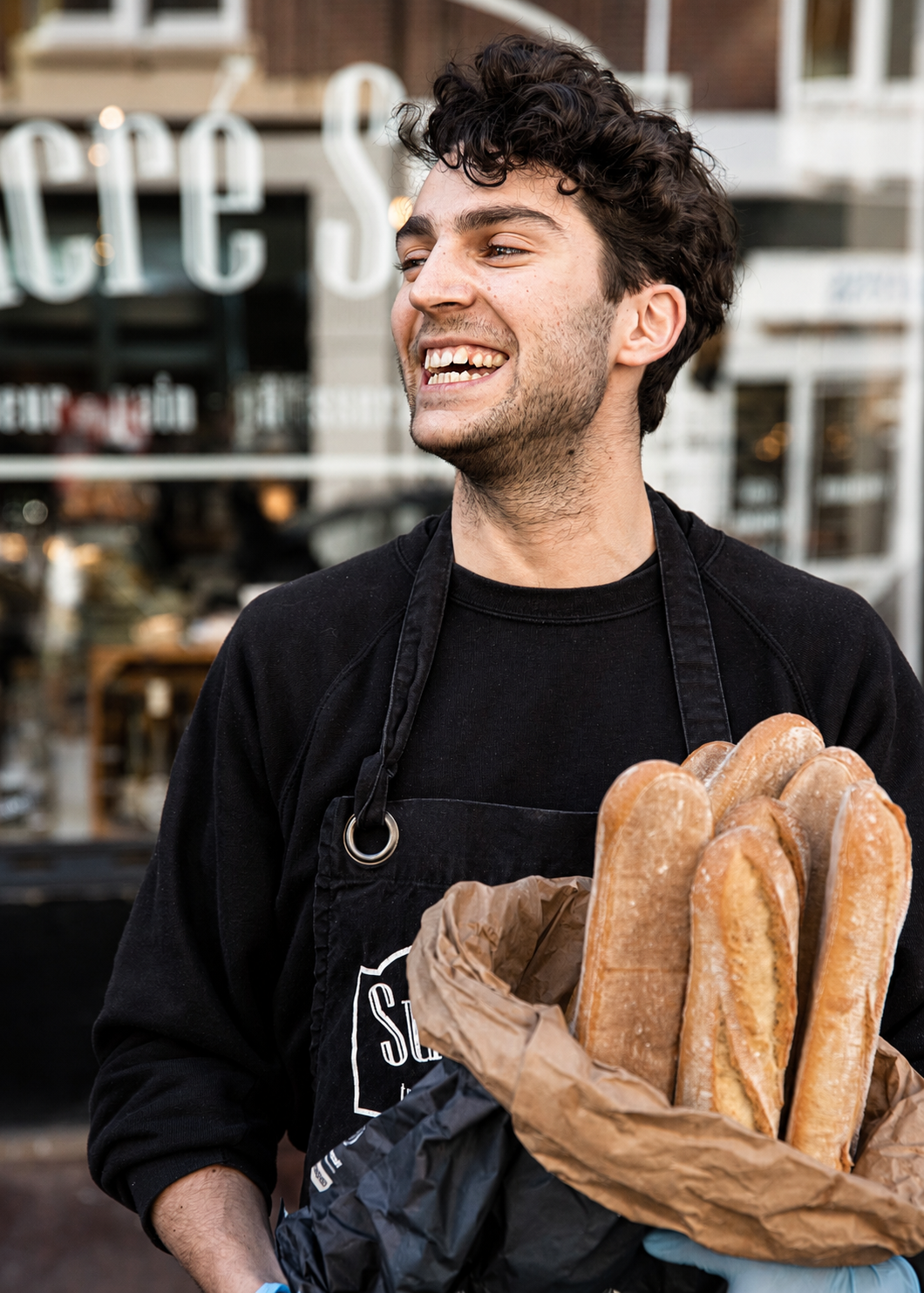 Young man smiling while holding a bunch of baguettes in a bakery or cafe setting.