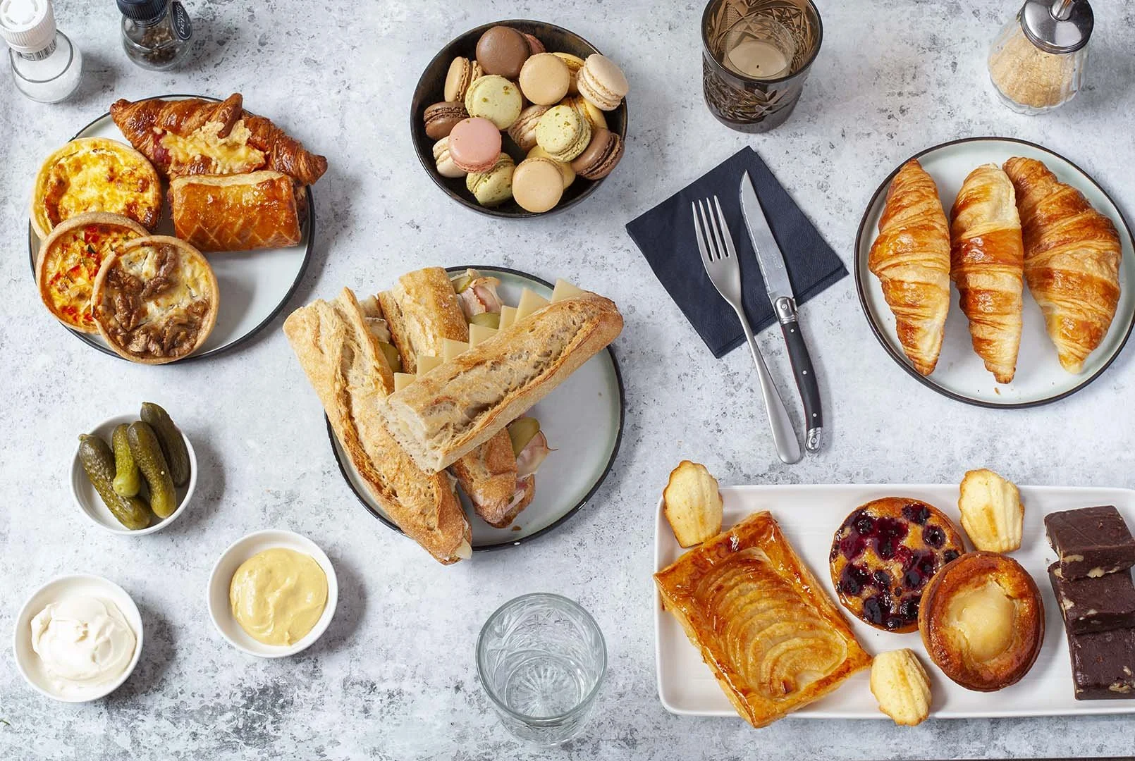 A breakfast spread with croissants, mini sandwiches, macarons, assorted sweet pastries, pickles, and condiments on a light textured table.