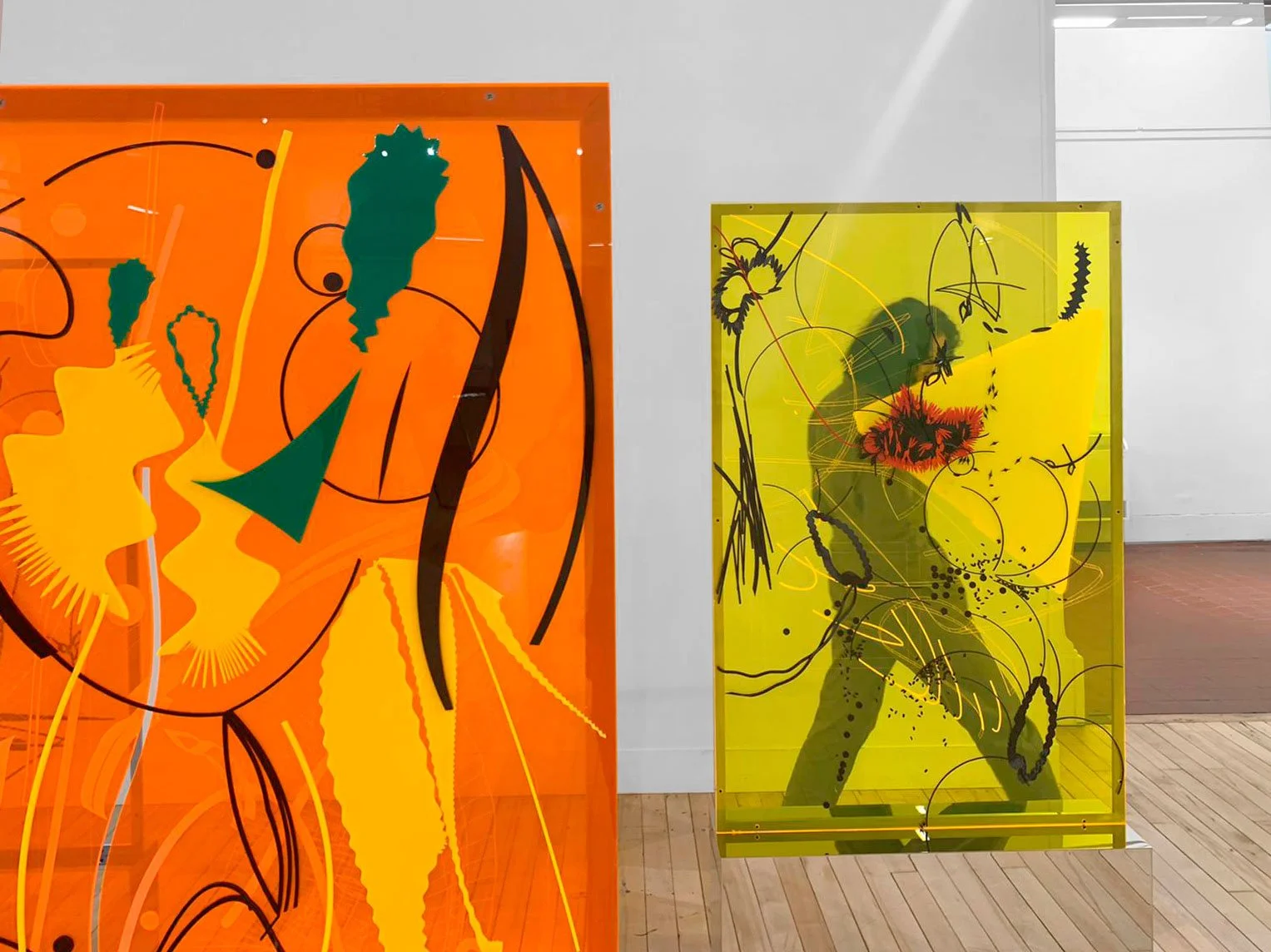 Two colorful abstract glass artworks, one orange and one yellow, featuring various shapes, lines, and patterns, displayed in an art gallery.