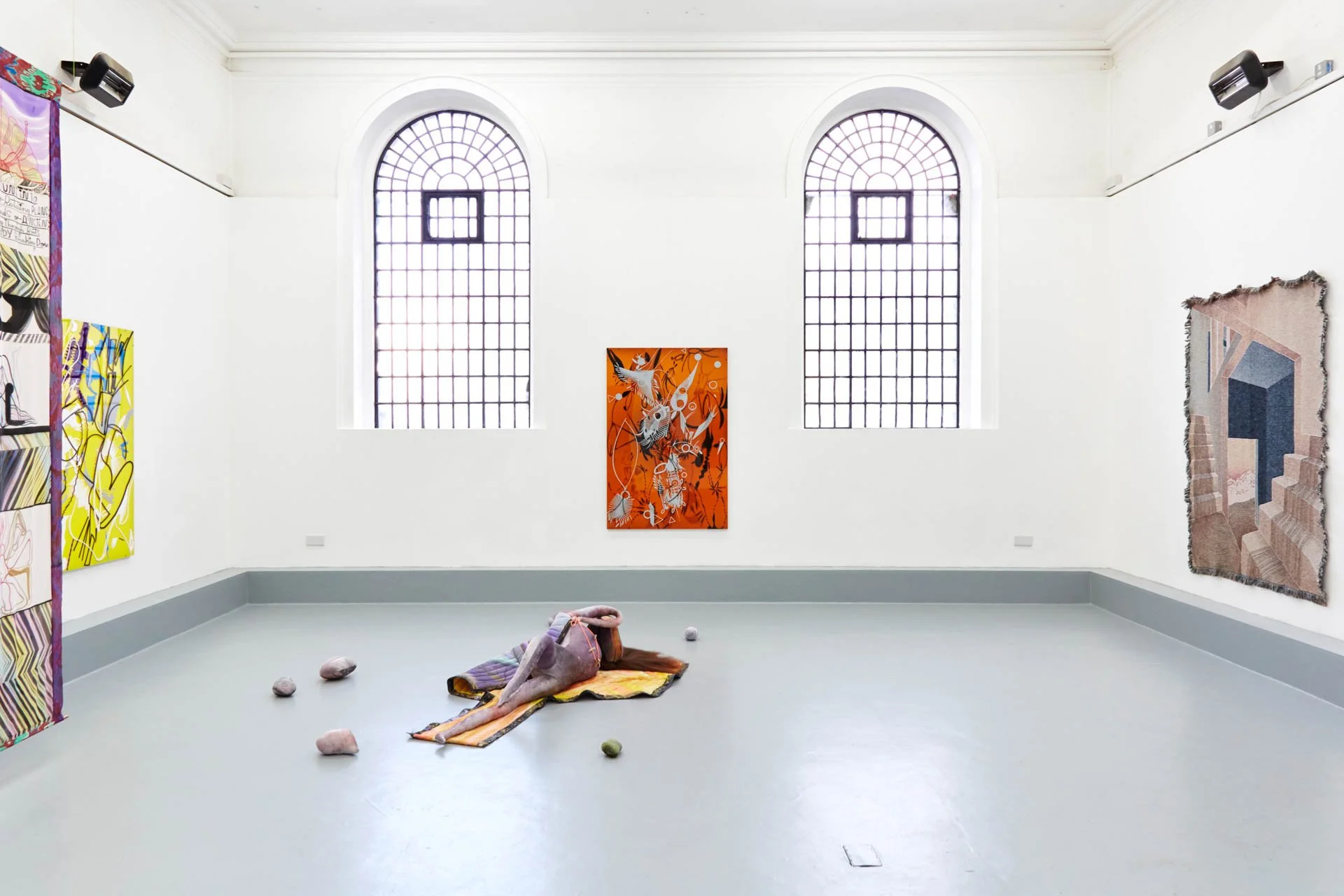 Art gallery with abstract paintings on white walls, featuring colorful paintings, a fabric sculpture on the floor, and large arched windows.