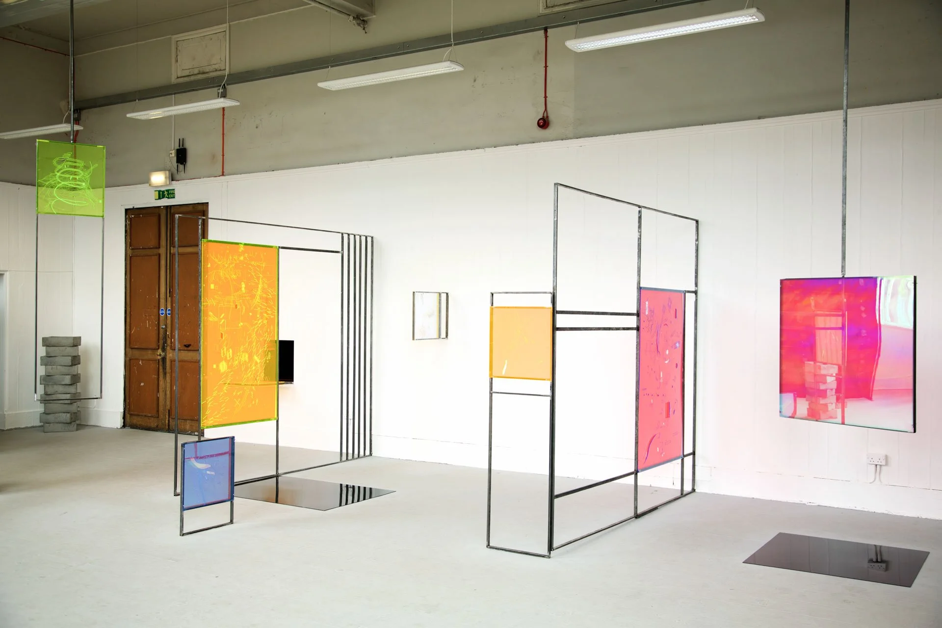Art installation in a gallery featuring colorful abstract digital displays on wire frames, with a white wall and ceiling lights in the background.