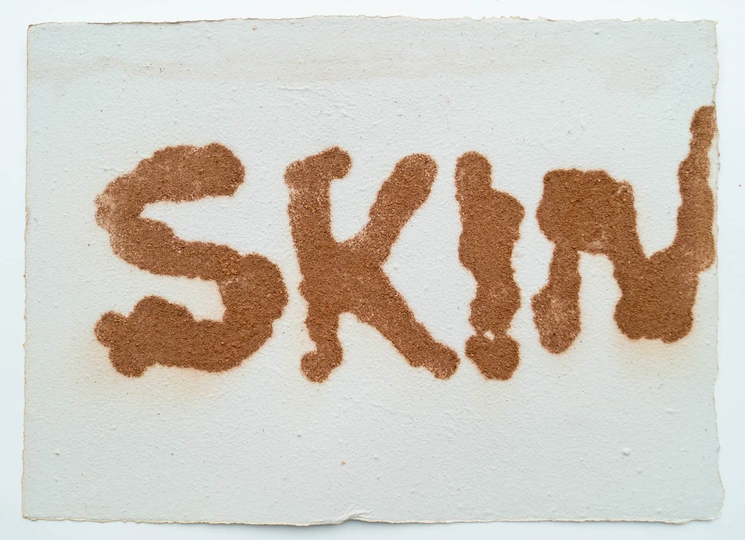 The word "SKIN" written with brown powder on a white textured paper.
