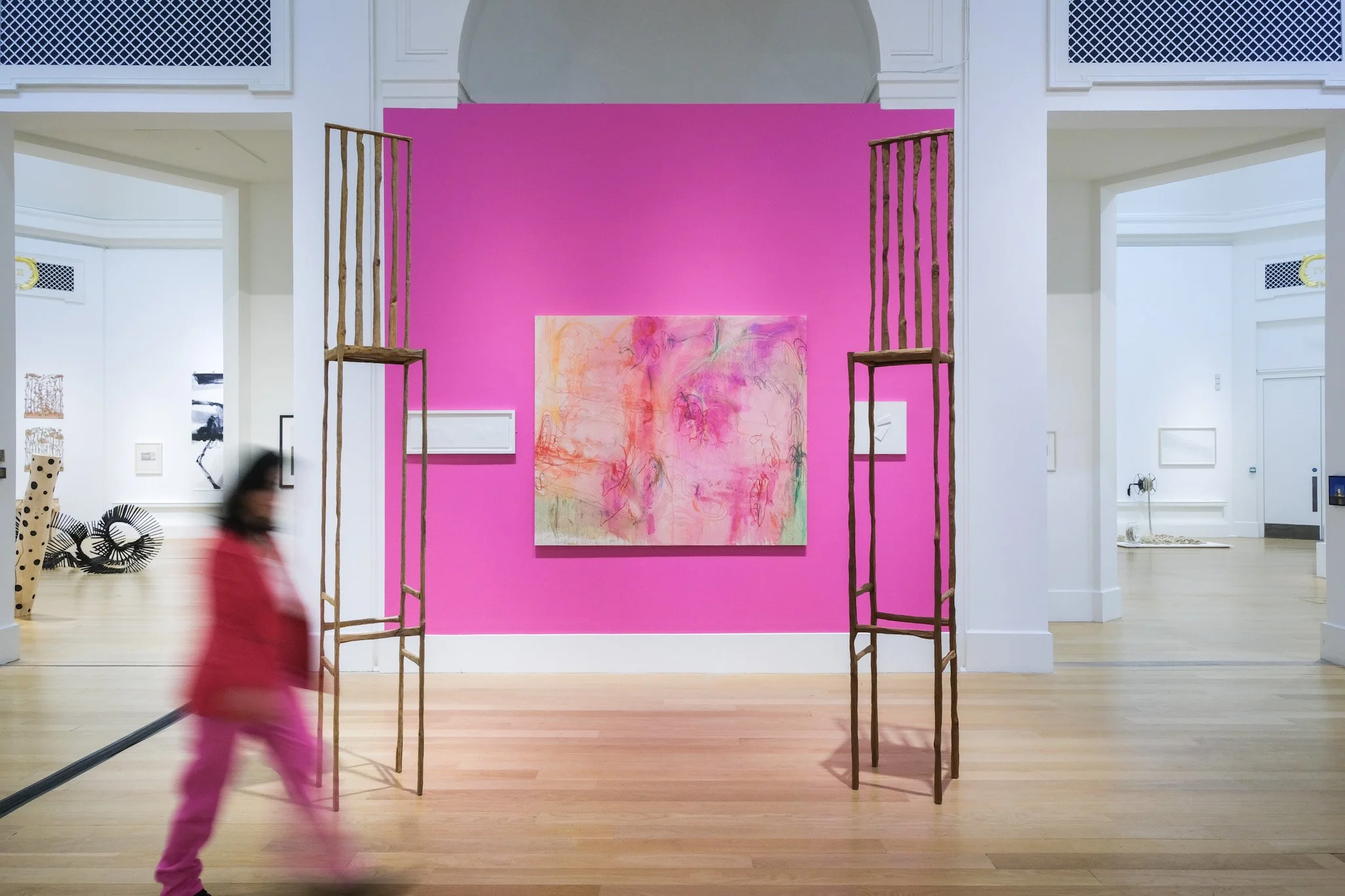 An art gallery with a pink wall featuring a colorful abstract painting, large wooden ladders on either side, and a blurred person walking in the foreground.