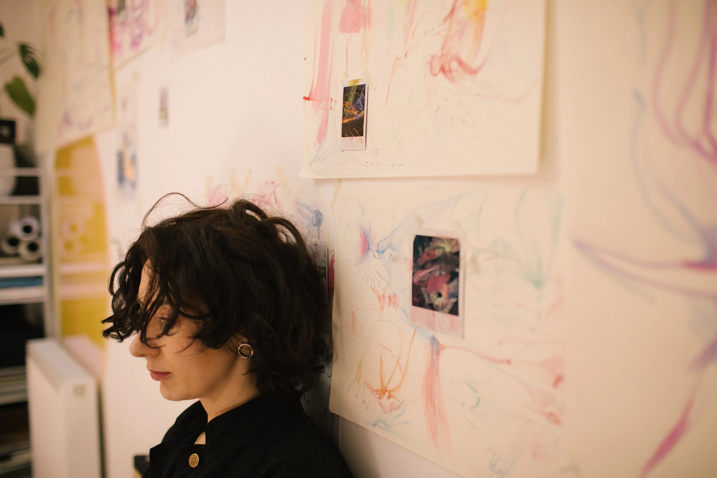 Woman with dark, curly hair and glasses leaning against a wall decorated with abstract colorful drawings and small photographs.