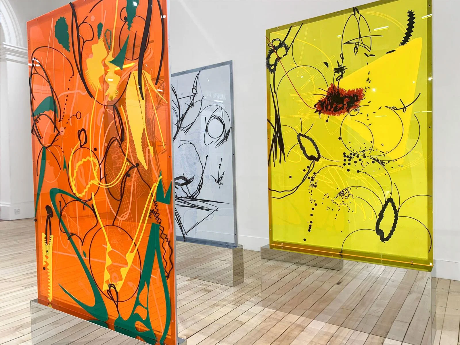 Three abstract art panels of glass with colorful, dynamic line designs, positioned in an art gallery with wooden floors and white walls.