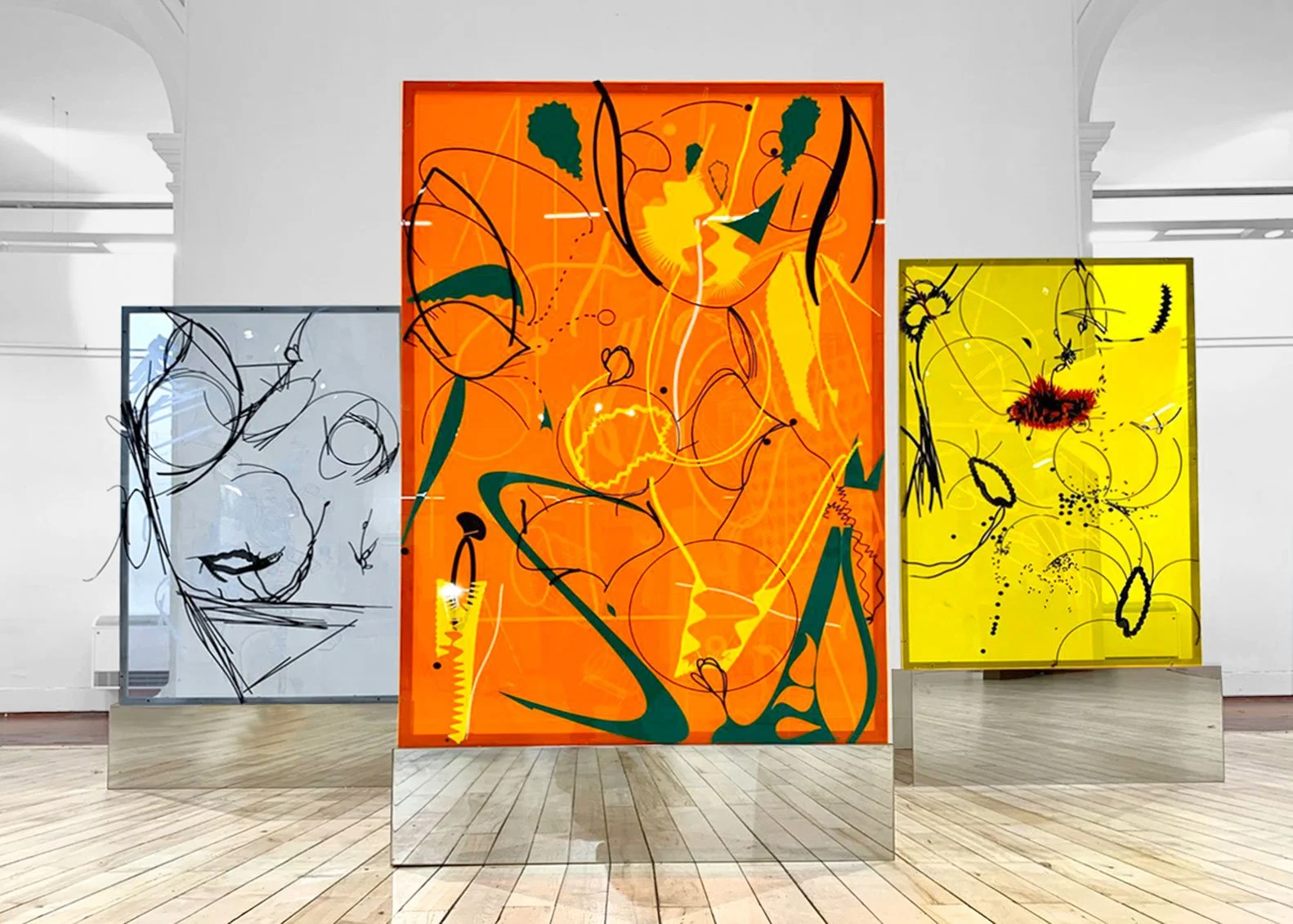 Three colorful abstract paintings on display in a gallery, with black, yellow, orange, and green lines and shapes.
