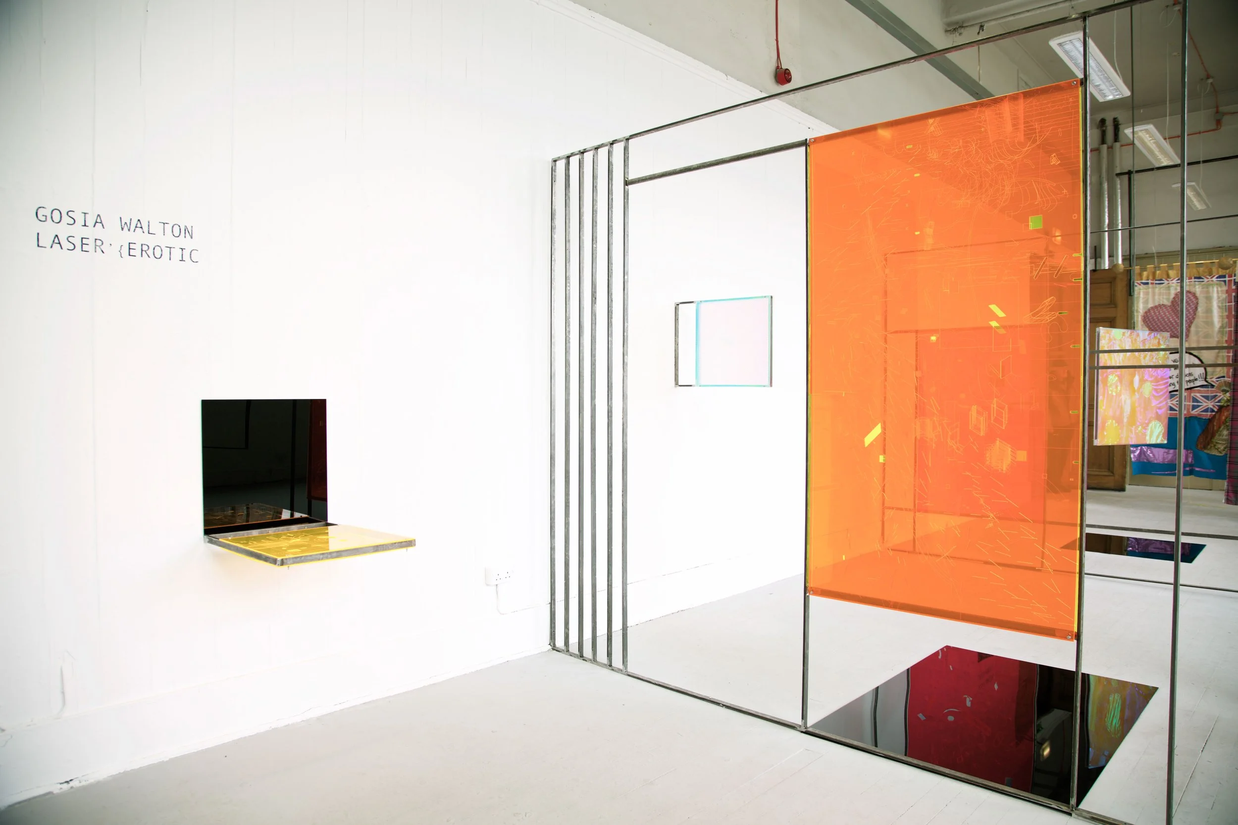 Contemporary art installation with a white wall, orange acrylic panel, metal frame, and a small square cutout window, titled 'GOSIA WALTON LASER {EROTIC}'.