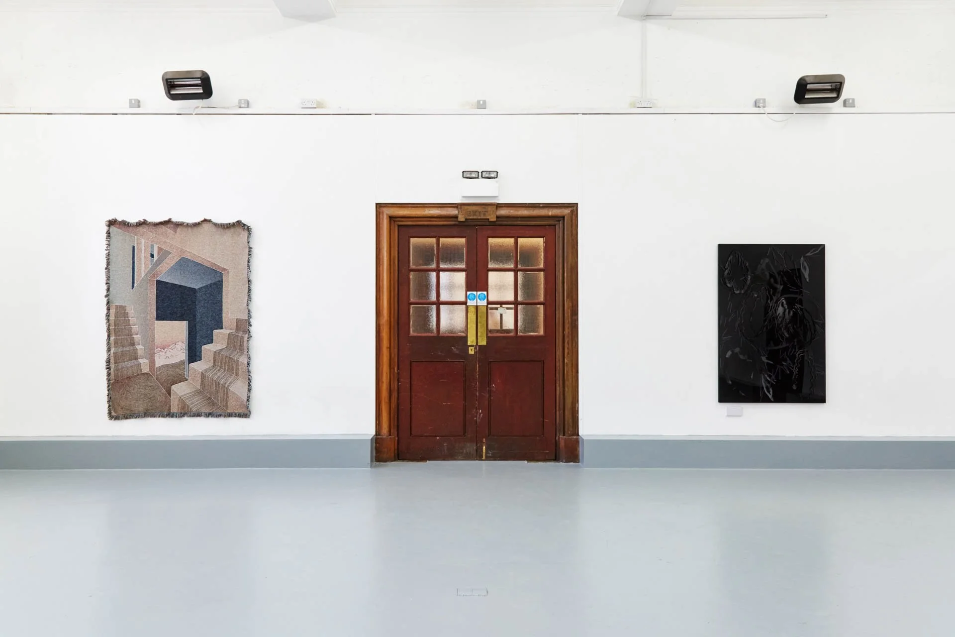 An art gallery with white walls and a wooden double door in the center, two artworks on the wall — a colorful woven or textile piece on the left, and a dark abstract painting on the right.