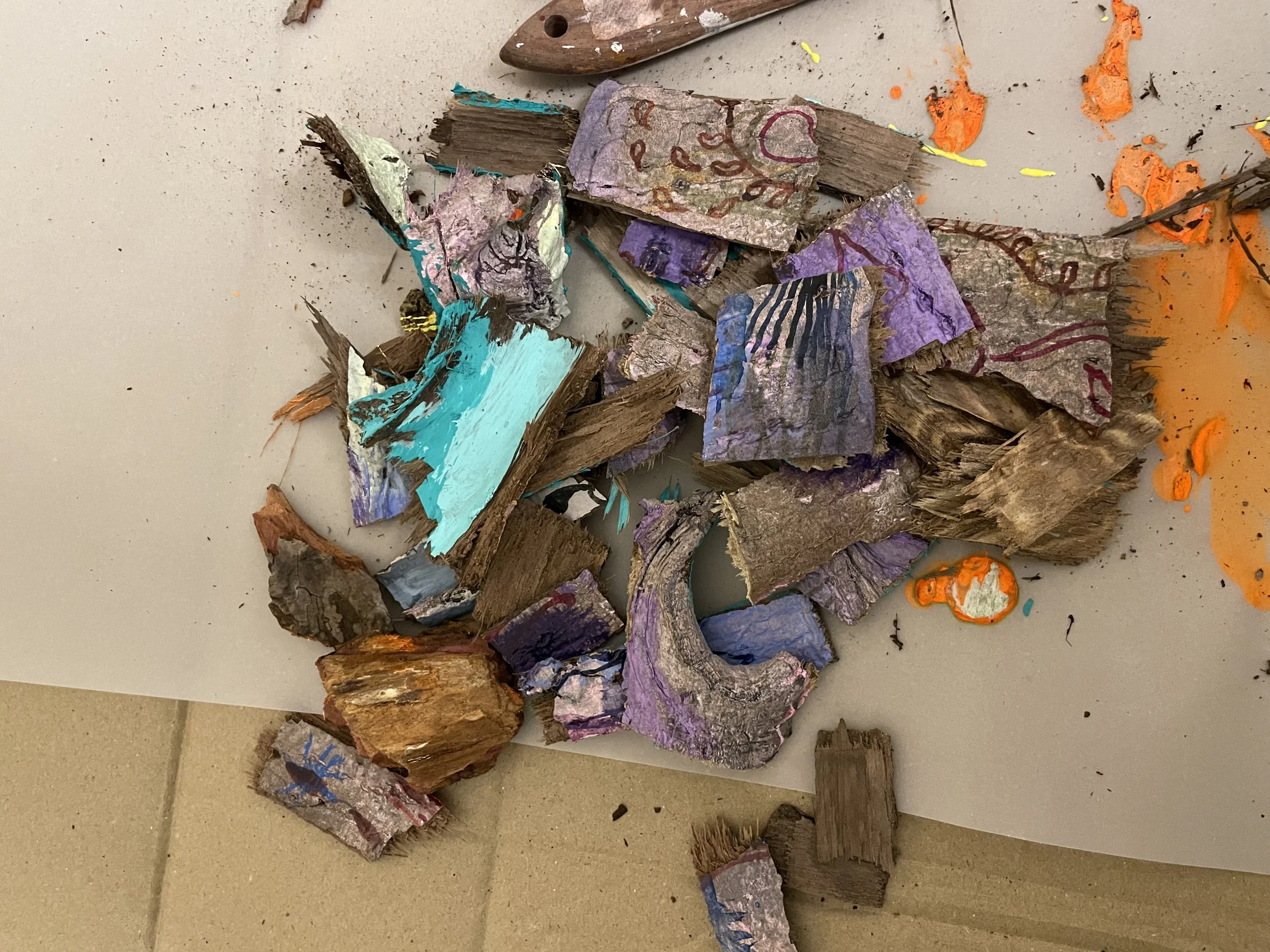 Broken pieces of paper with colorful drawings and paint, some wood chips, and a plastic palette knife on a beige surface.