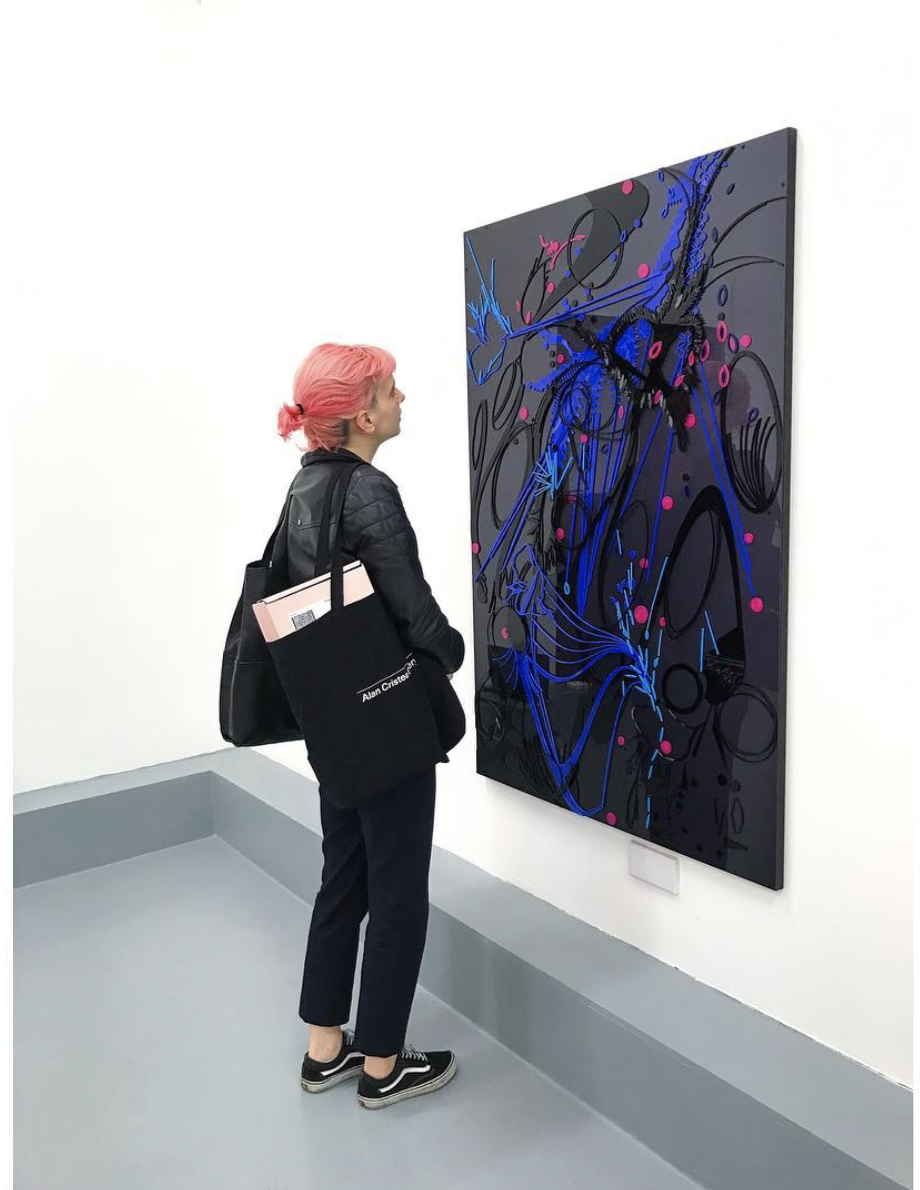 A woman with pink hair, wearing a black jacket and dark pants, is standing in an art gallery, looking at an abstract painting with black, blue, and pink elements on a black canvas.