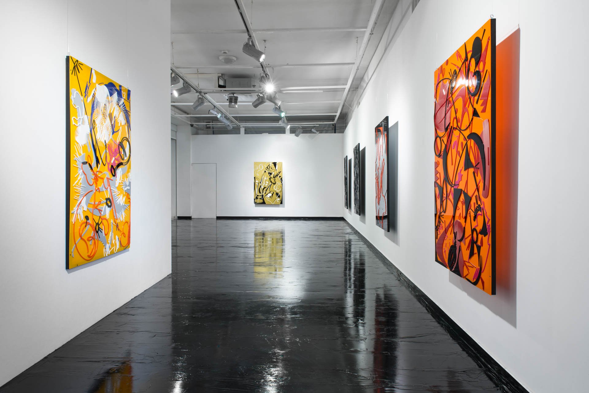An art gallery with white walls and black flooring, displaying colorful, abstract paintings. Track lighting illuminates the artwork, with several paintings on the left and right walls and one on the back wall.