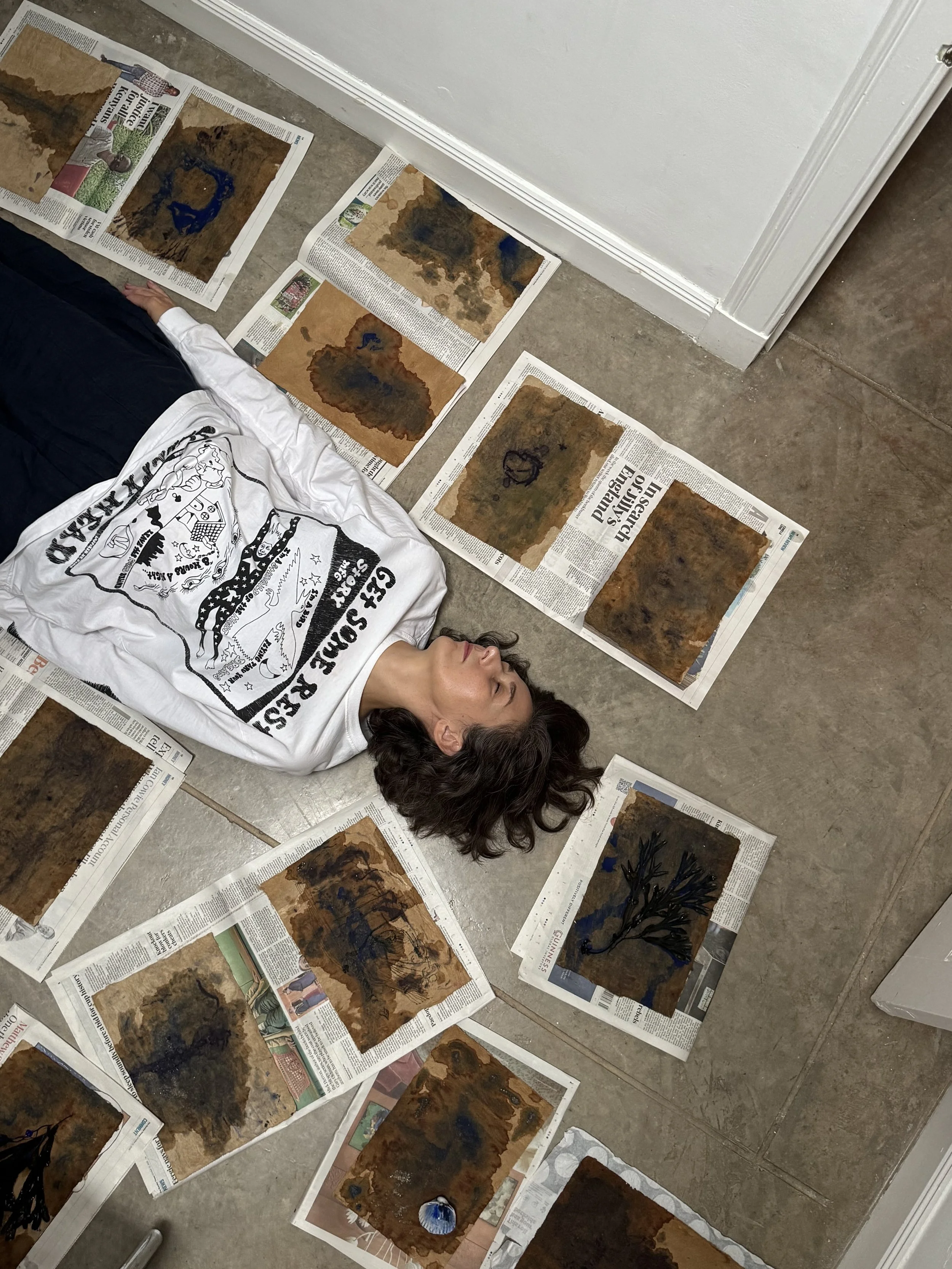 Person lying on the floor surrounded by watercolor paintings on newspapers.