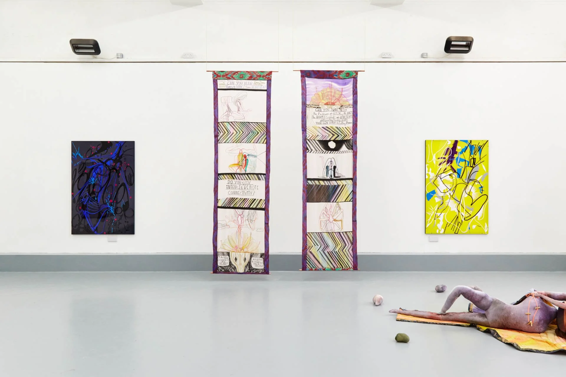Art gallery with three colorful abstract paintings on a white wall and a textured sculpture of a reclining figure on the floor.