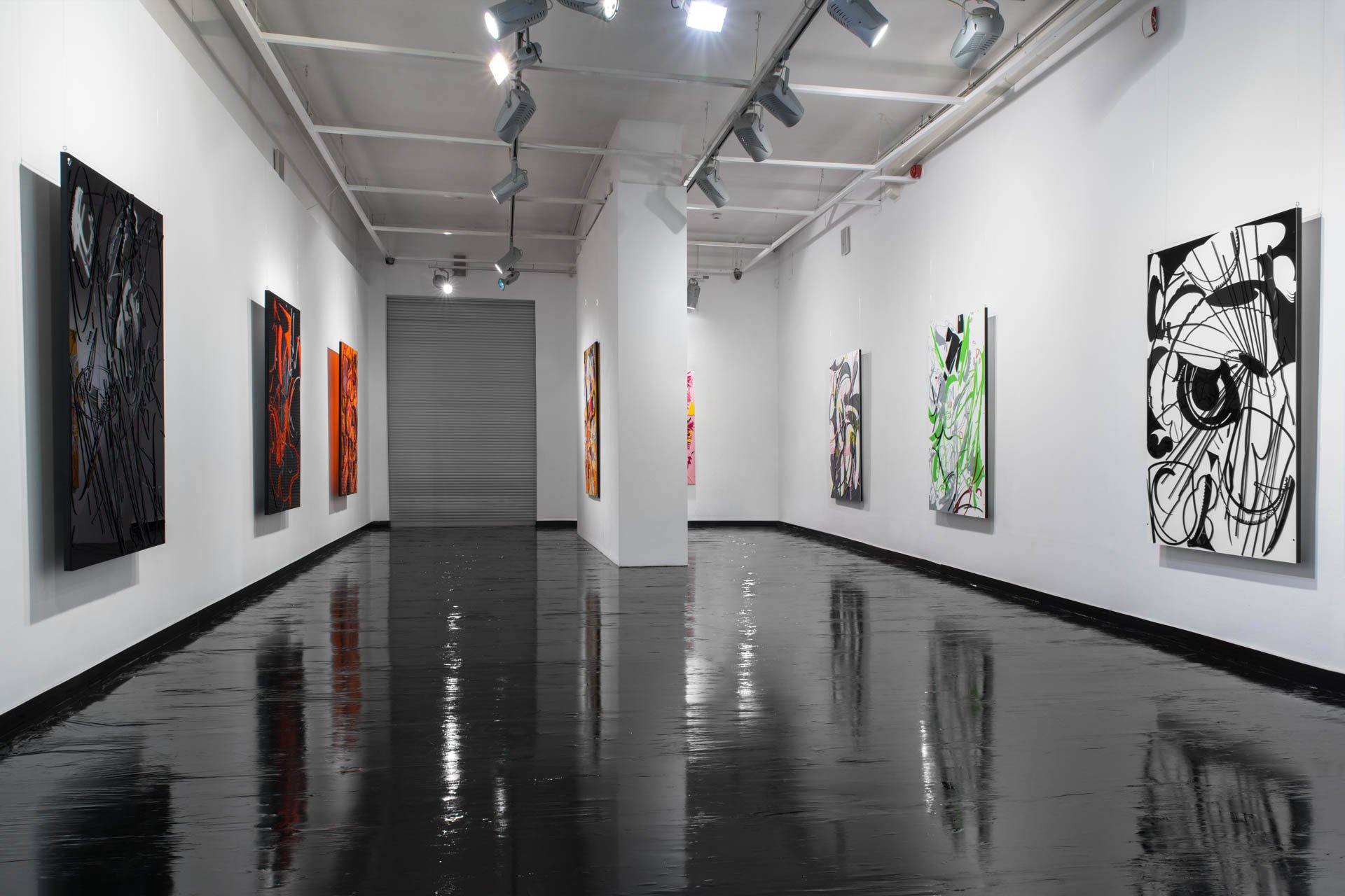 An art gallery with white walls, black glossy flooring, and colorful abstract paintings displayed on the walls. Overhead lighting illuminates the artwork.