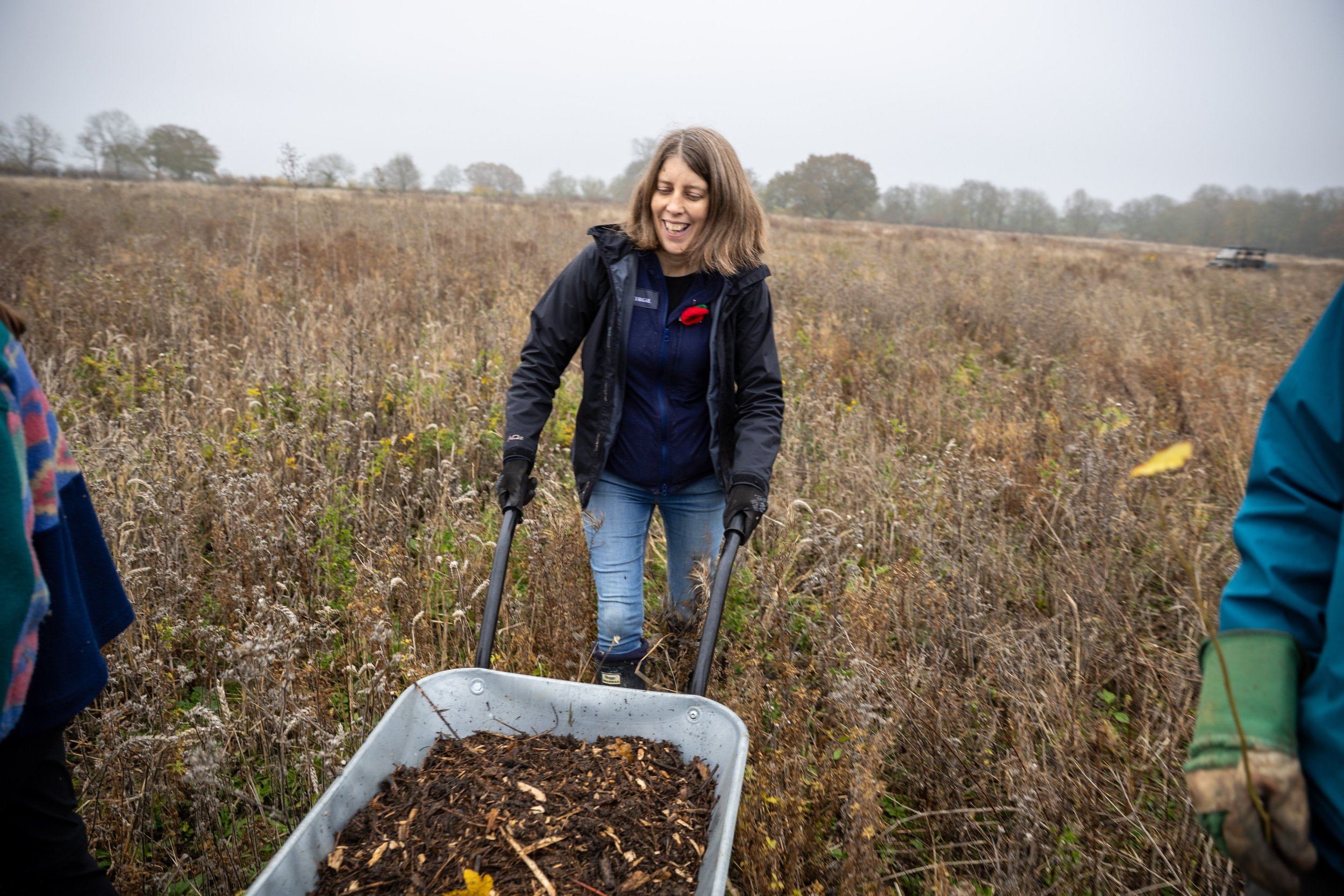 Woman in outdoor gear pushing a wheelbarrow filled with soil through a field of dry plants