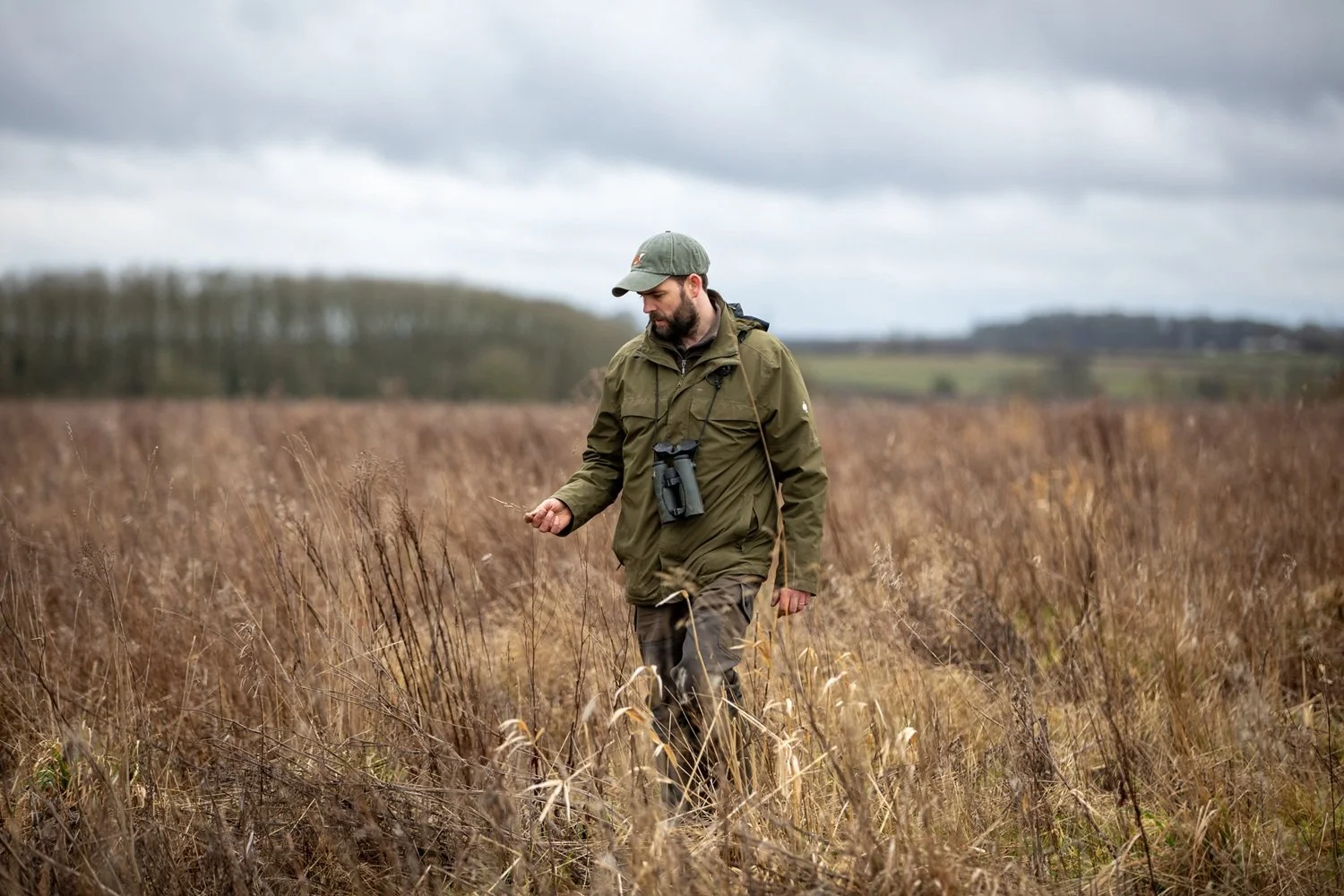 A man walking through a field of tall, dry grass, wearing outdoor clothing, a baseball cap, and binoculars around his neck, under a cloudy sky.