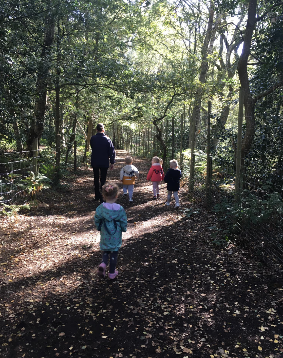 A group of five children and one adult walking on a dirt path through a wooded area with sunlight filtering through the trees.
