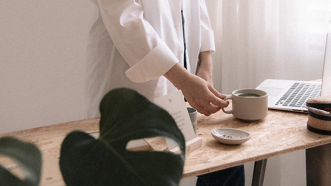 Hyvän elämän valmentaja Terhi Lahti työpöydän ääressä. Hän pitää kädellään teekupista kiinni. Pöydällä läppäri ja muuta pientavaraa. Edustalla viherkasvi.