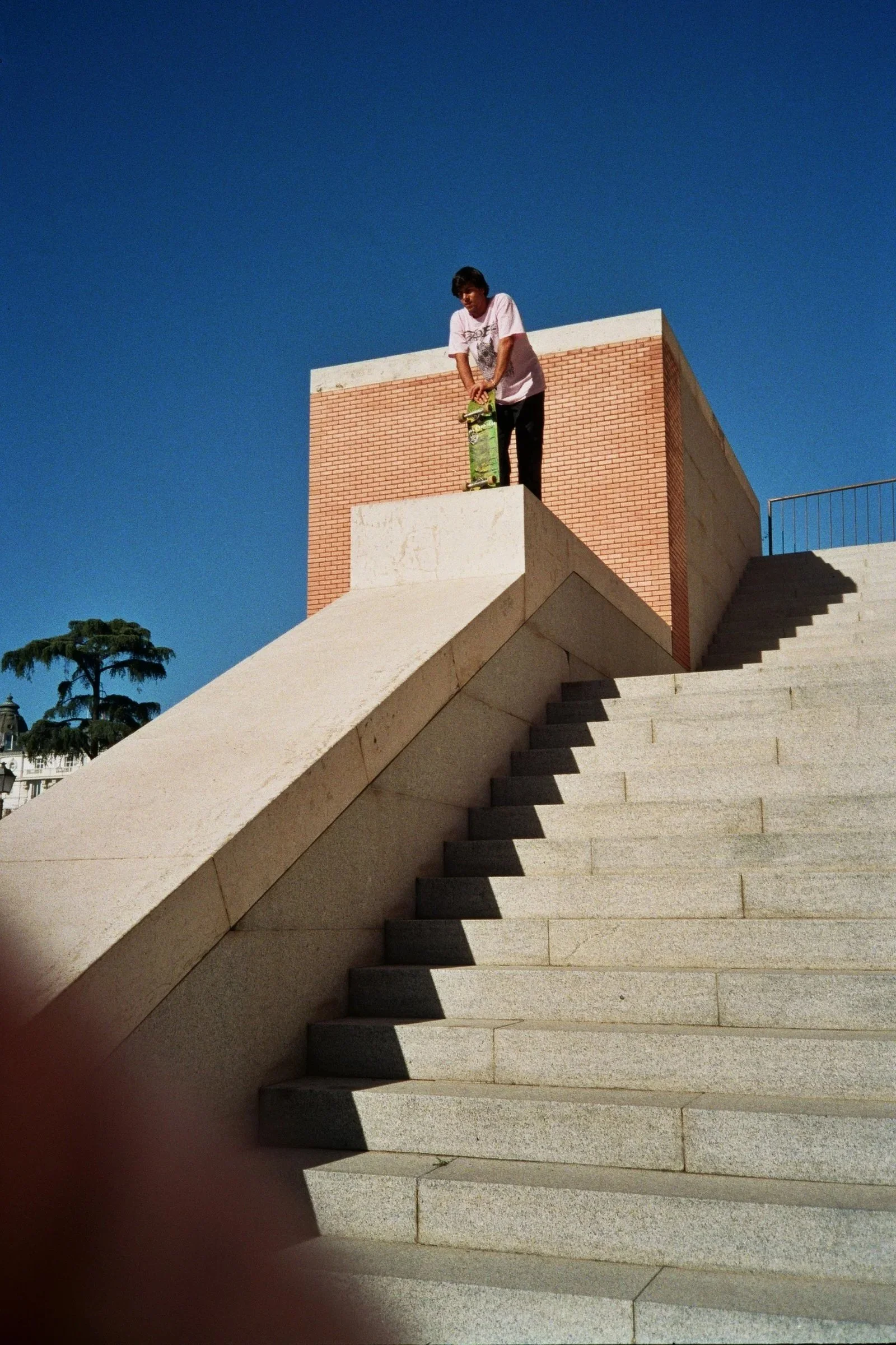 Joven en una escalinata de piedra, en una comunidad urbana, preparando para hacer trucos con una patineta, con un edificio de ladrillos y cielo despejado en el fondo.