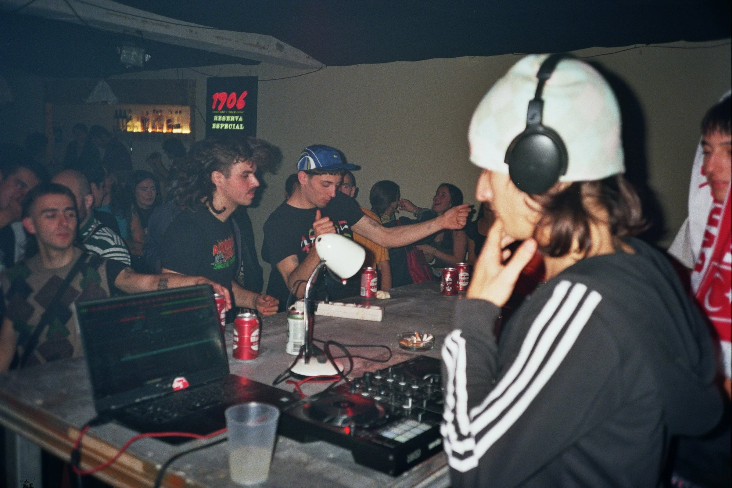 Personas en una fiesta con DJ, computadora portátil, mezcladora y bebidas en la barra.