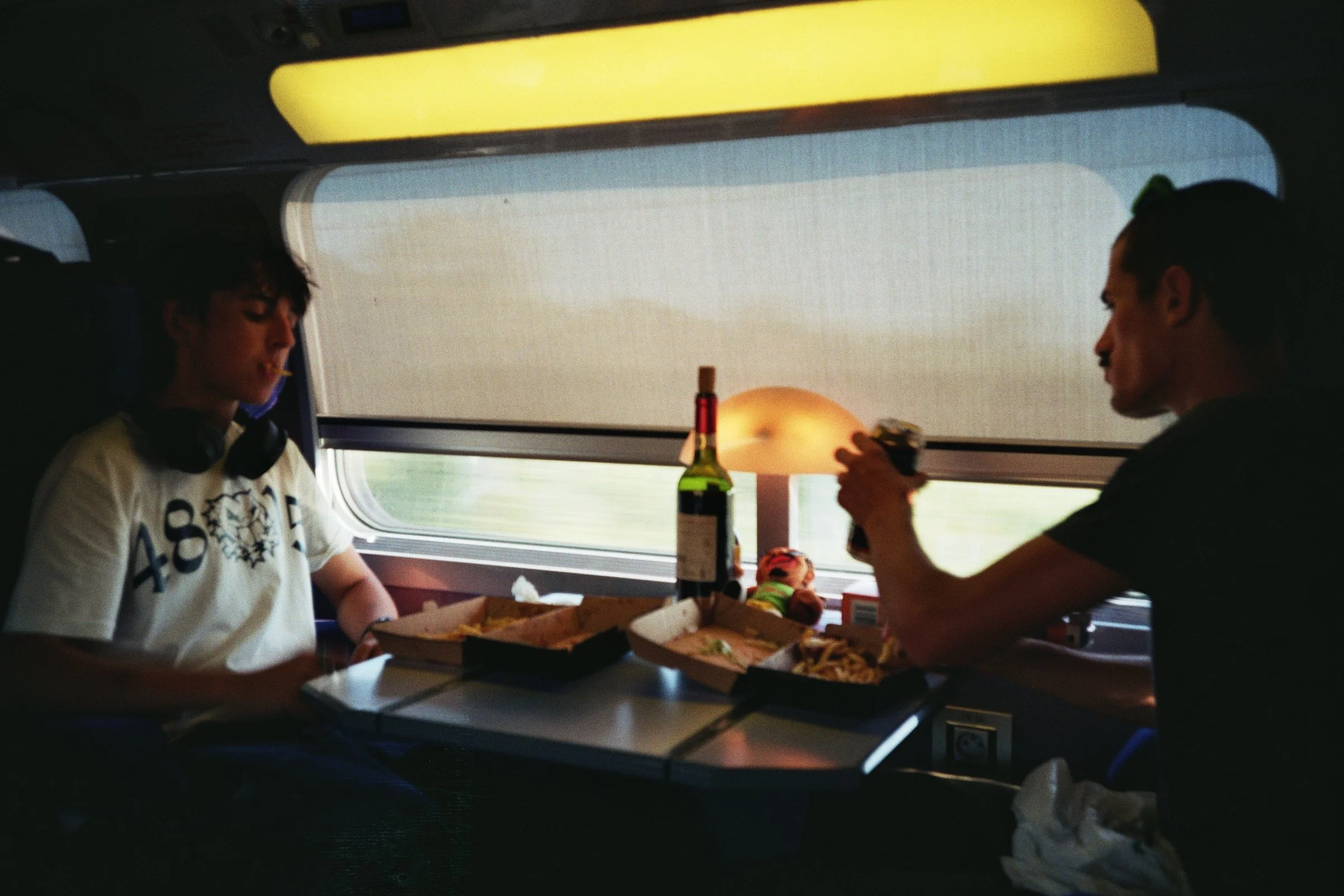Dos personas cenando en un tren, una joven con auriculares y camiseta blanca, y un adulto con camiseta negra, con comida en cajas para llevar y una botella de vino en la mesa.
