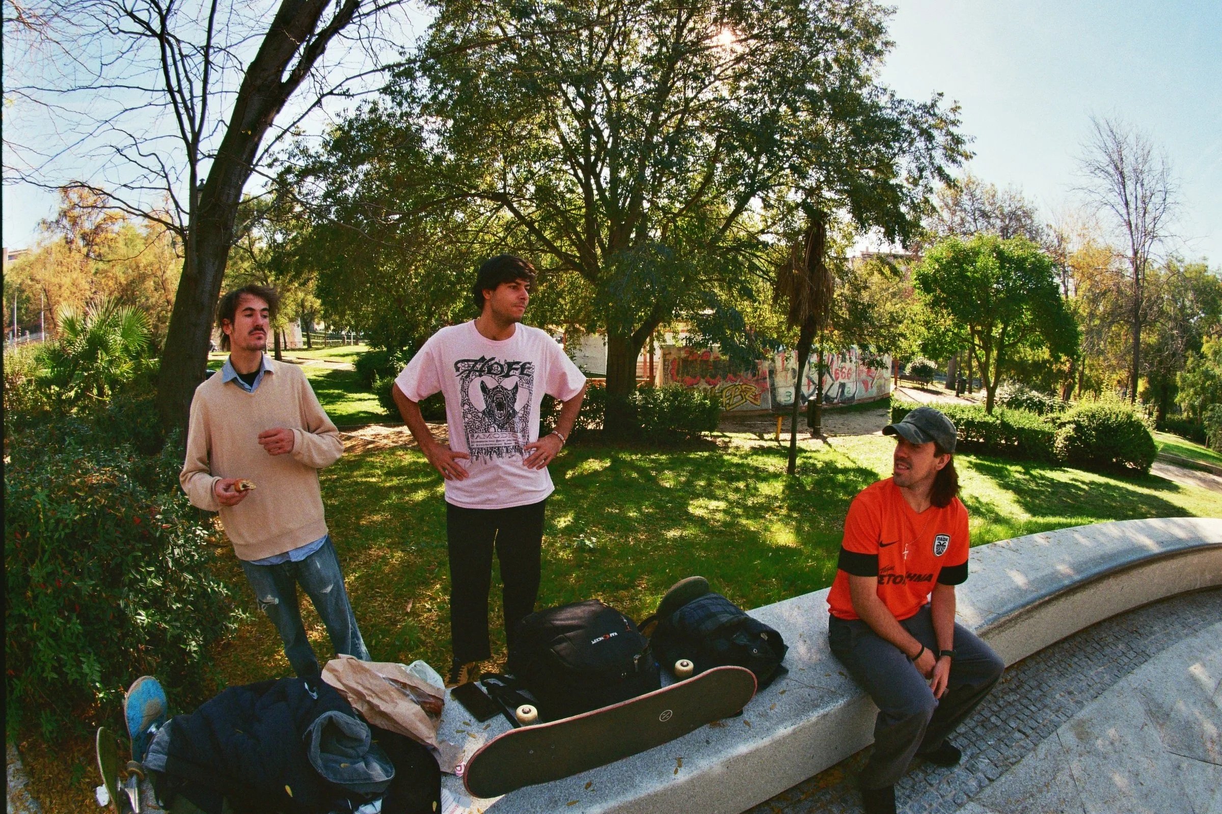 Tres jóvenes en un parque con árboles verdes, uno está sentado en un banco de cemento con una camiseta naranja y gorra, los otros dos están de pie, uno con suéter y el otro con camiseta blanca con diseño, rodeados de mochilas y patineta.
