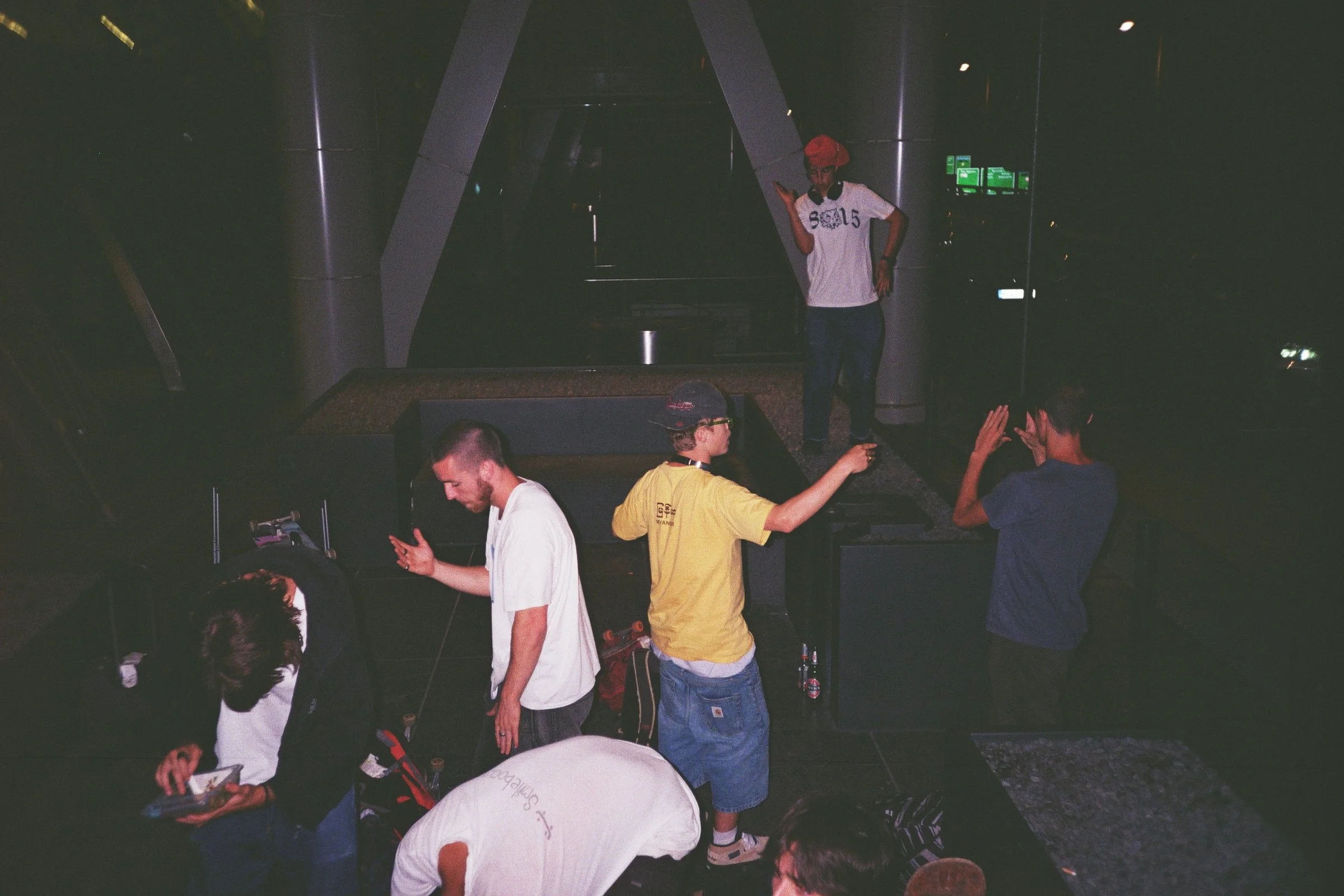 Grupo de jóvenes en un espacio oscuro, algunos viendo sus teléfonos y otros posando o interactuando en un ambiente que parece una sala o un lugar de actividades.