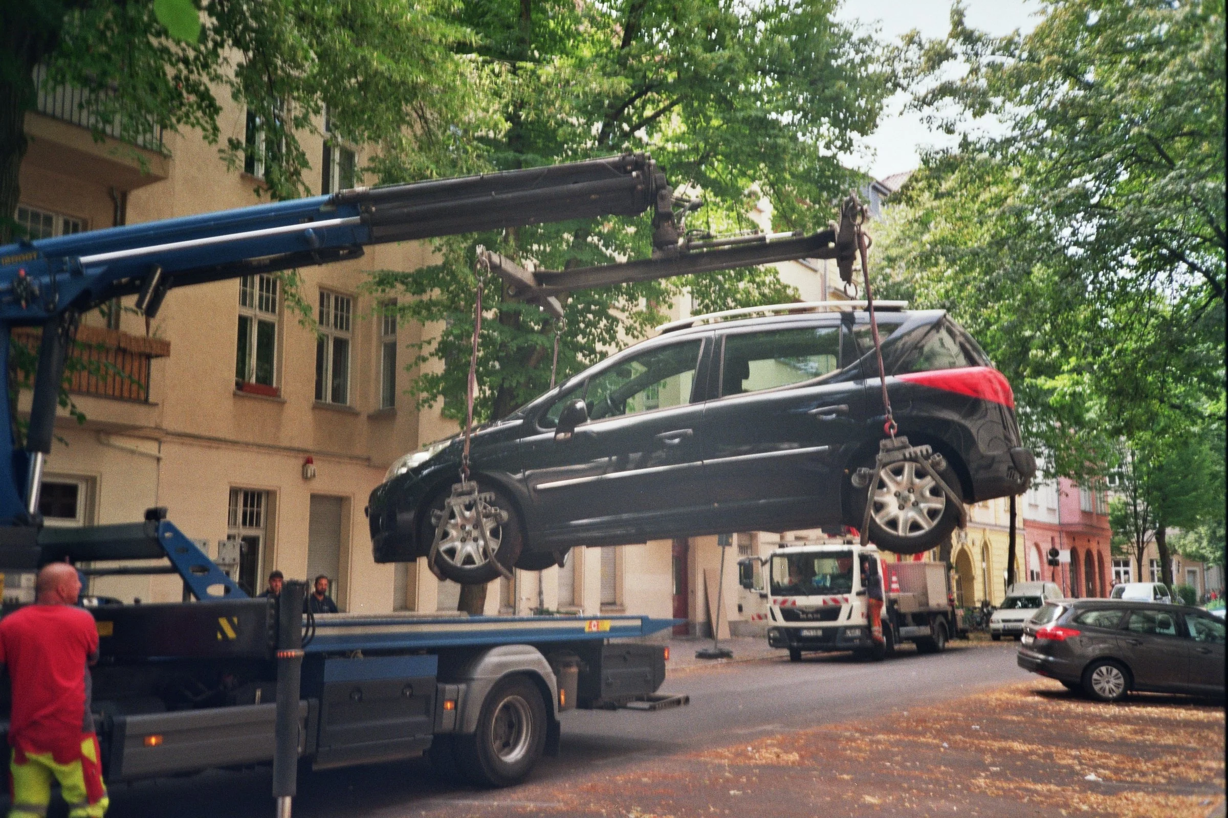 Un automóvil negro está siendo levantado por una grúa en una calle con árboles y edificios residenciales. Hay varios vehículos estacionados y dos personas observando la situación.