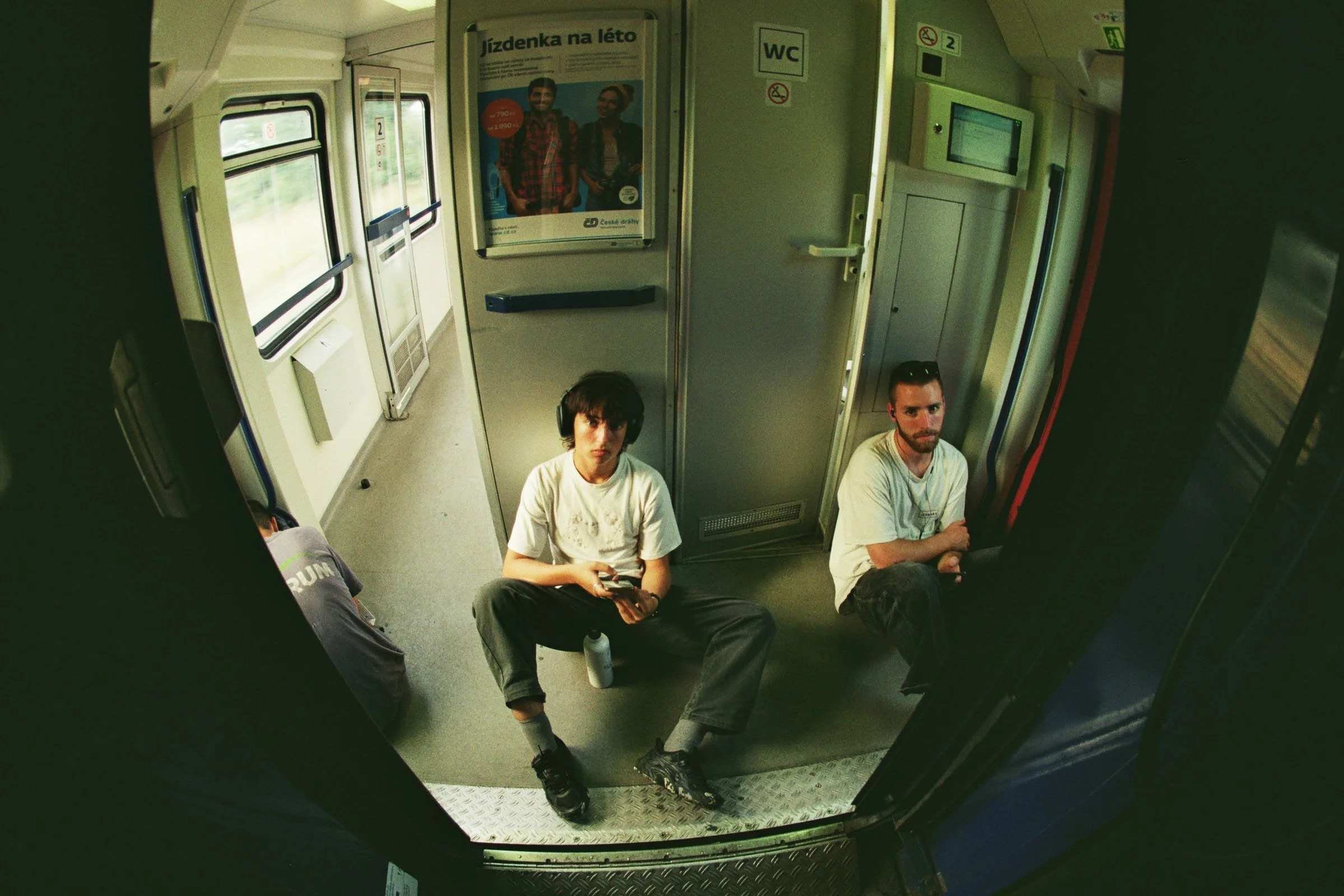 Resultados de una cámara en el interior de un tren, mostrando a dos hombres sentados en el suelo cerca de la puerta, uno con auriculares y el otro con barba, con ventanas y anuncios en las paredes del tren.