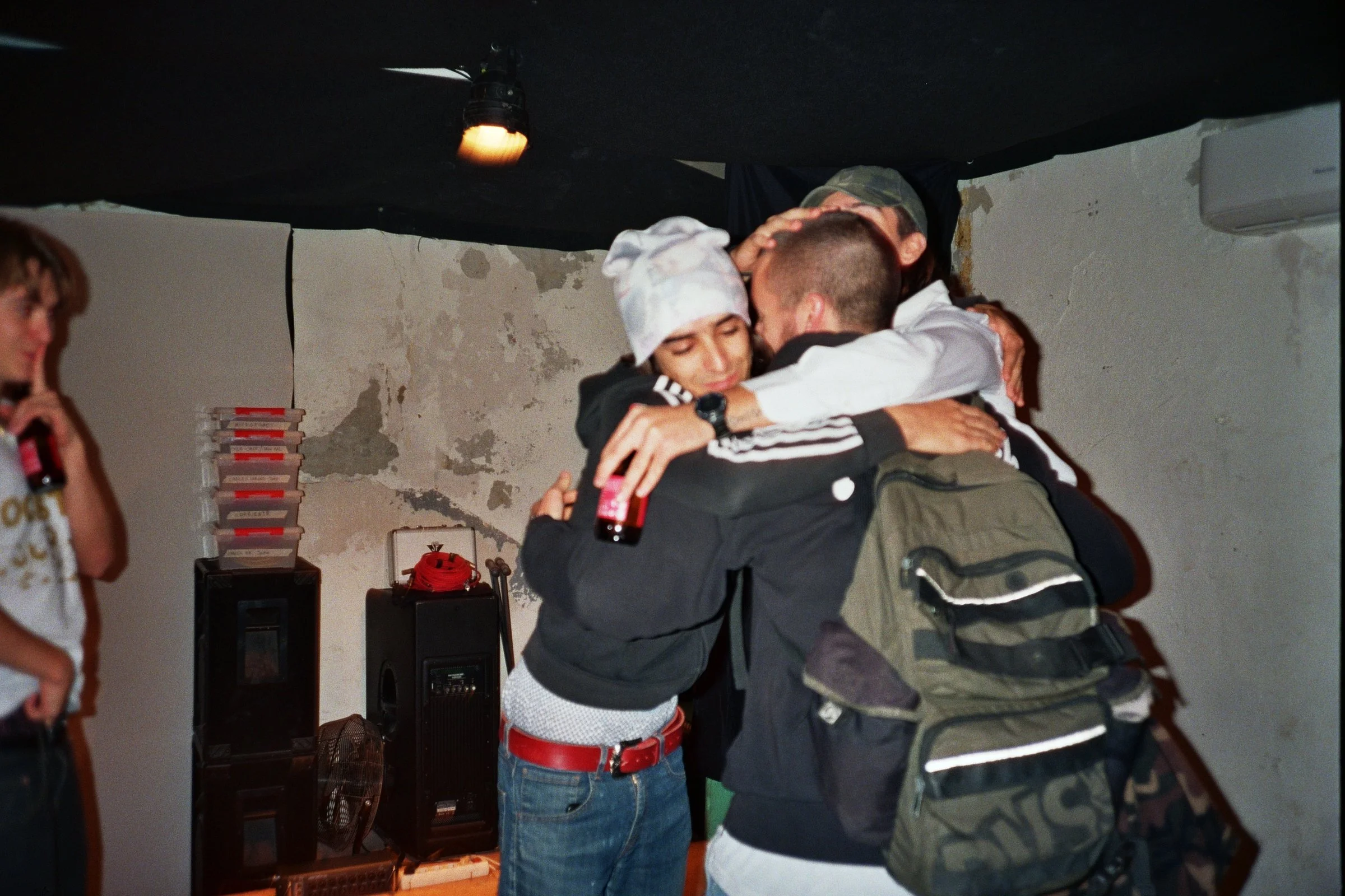 Tres jóvenes en una fiesta se abrazan con alegría en un ambiente con paredes deterioradas, rodeados de equipo de sonido y un ventilador.