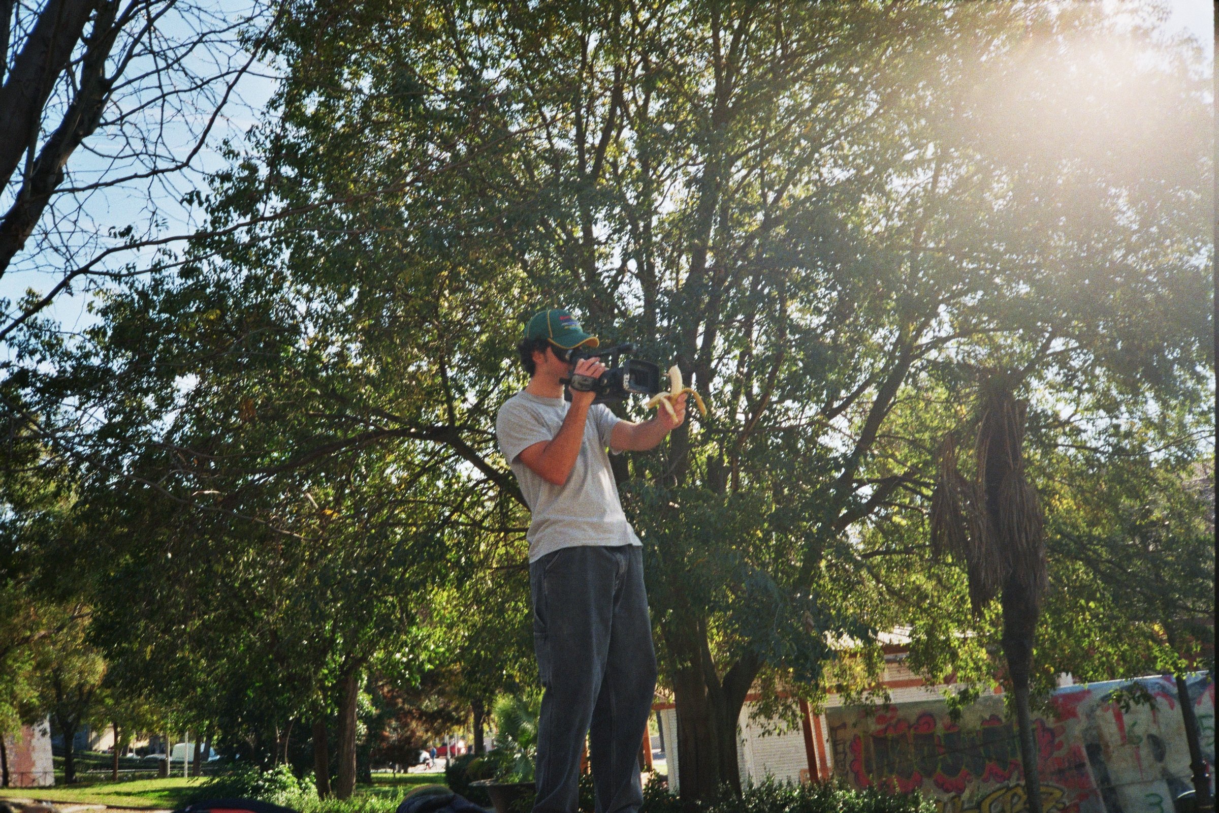Un joven con gorra y camiseta gris, sosteniendo una cámara de video, en un parque con árboles y graffiti en la pared, mientras toma una banana.